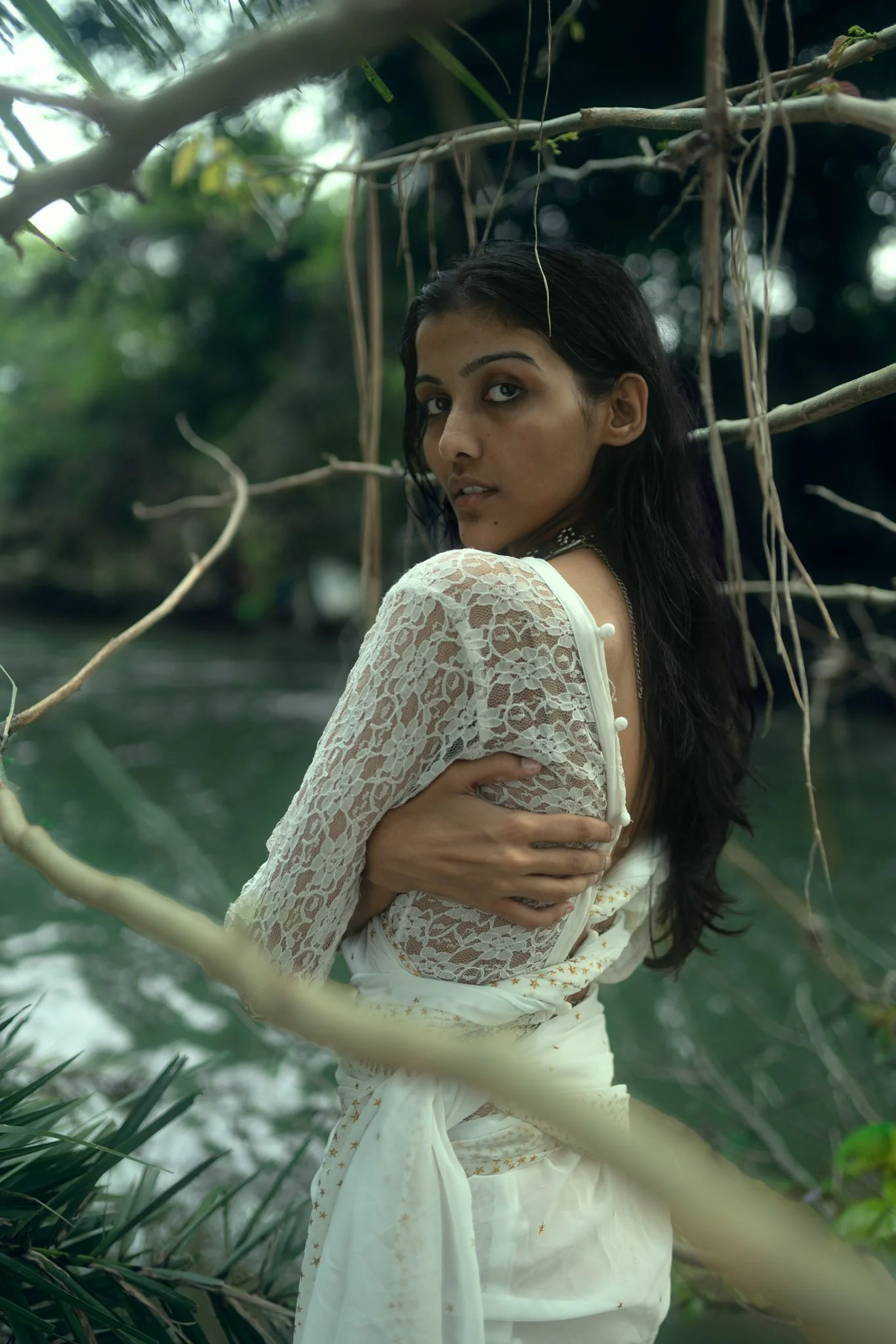 A woman in a white lace dress standing near a body of water, surrounded by tree branches and greenery, looking over her shoulder.