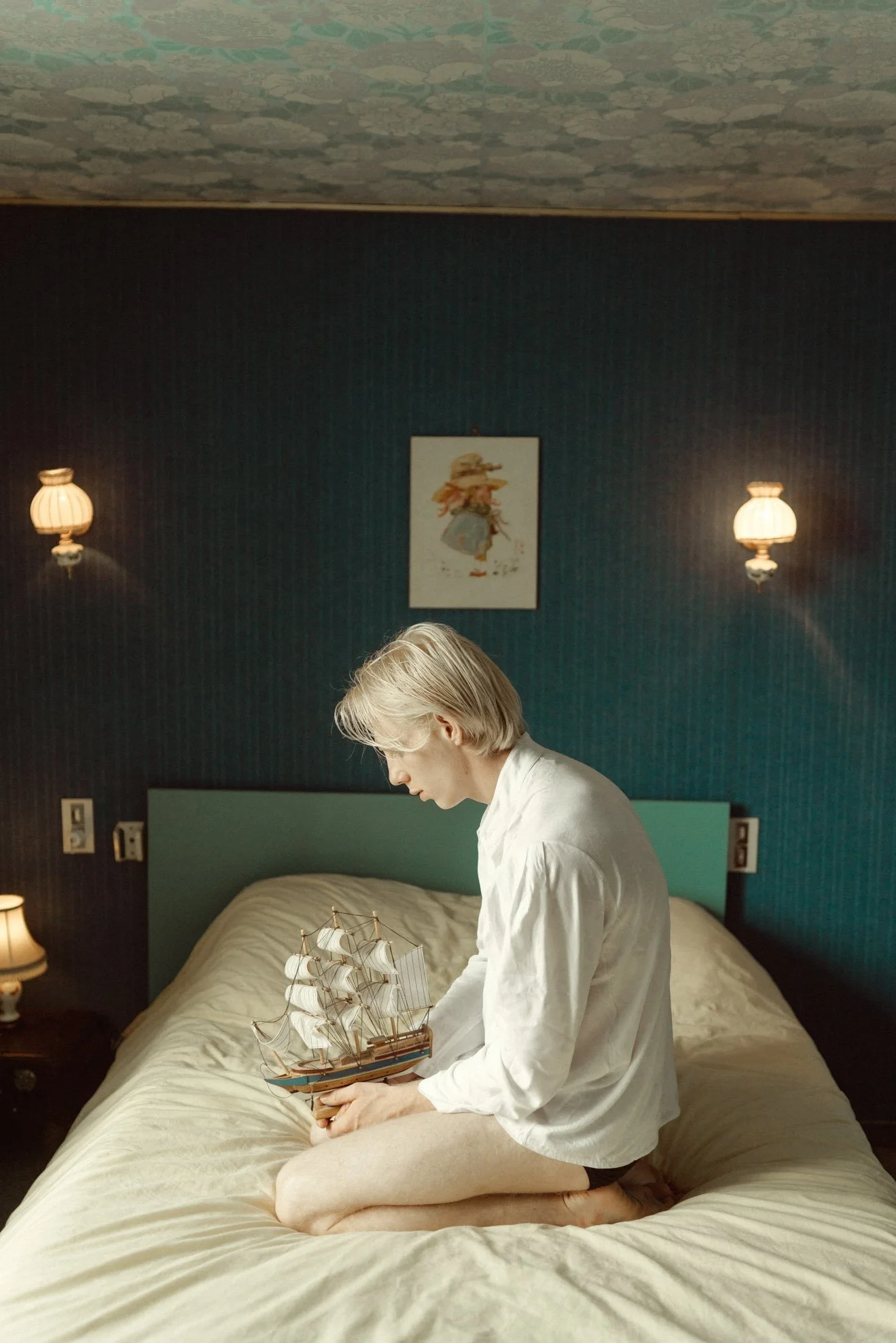 A young man with blonde hair sitting on a bed, holding a model sailing ship, in a dimly lit room with teal walls, two wall lamps, and a framed picture above the bed.