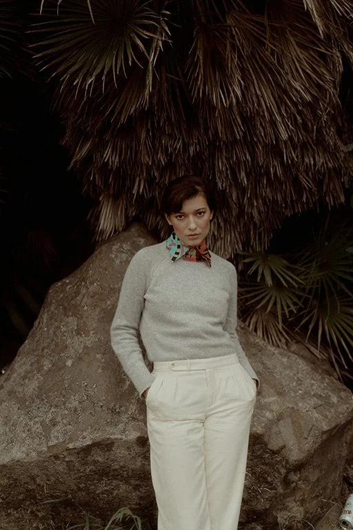 A woman stands outdoors in front of a large rock and tropical plants, wearing a gray sweater, white pants, and a colorful patterned scarf around her neck.