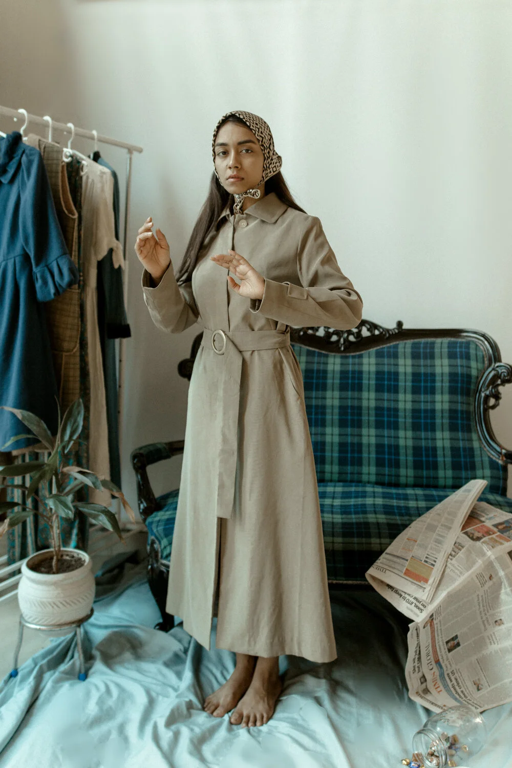 A woman standing on a bed in a room with a clothing rack, a plaid sofa, a potted plant, and newspapers, wearing a beige trench coat and a patterned headscarf.
