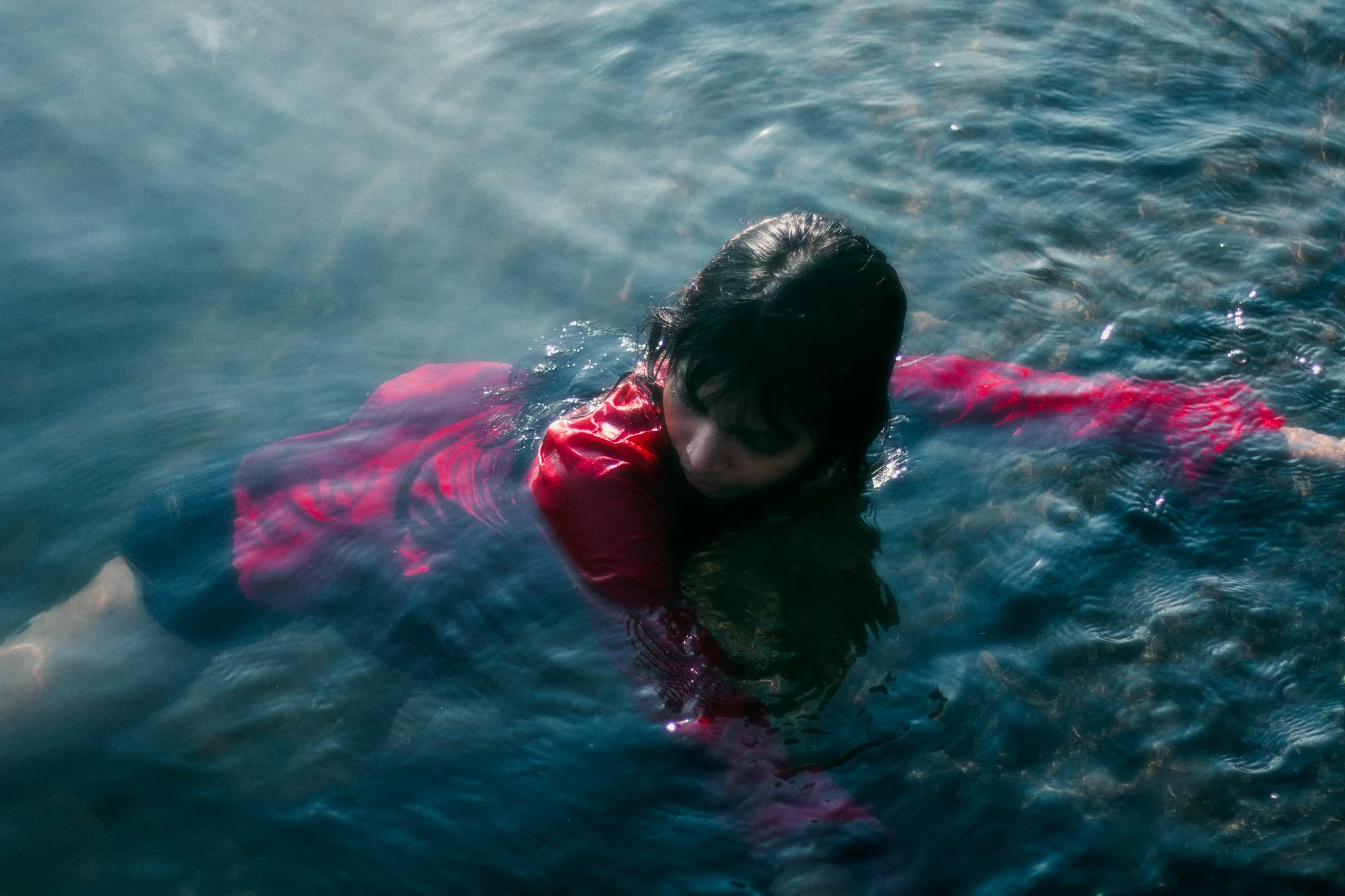 A woman in a red jacket is lying face down in the water, partially submerged, with her wet hair covering part of her face.