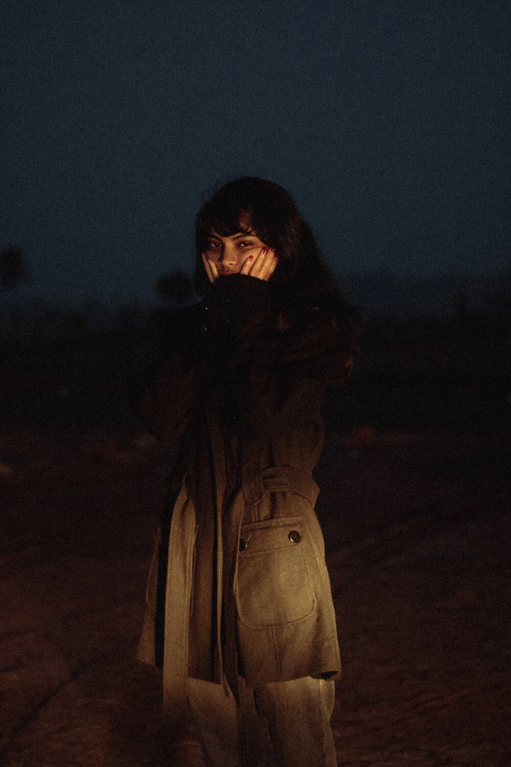 A woman standing outdoors at night, covering her face with her hands and smiling.