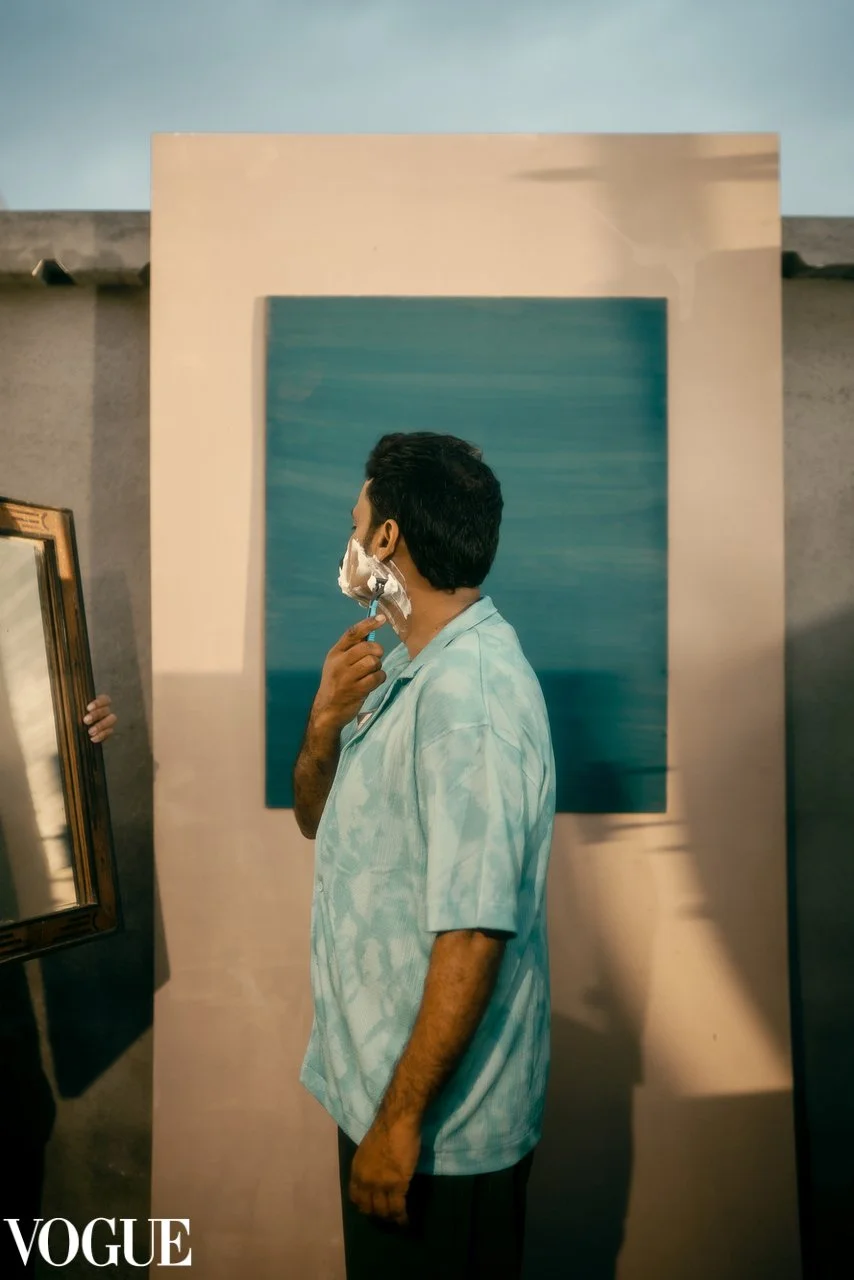Man with shaving cream on his face, standing in front of a blue abstract painting, holding a mirror in his left hand at an art gallery.