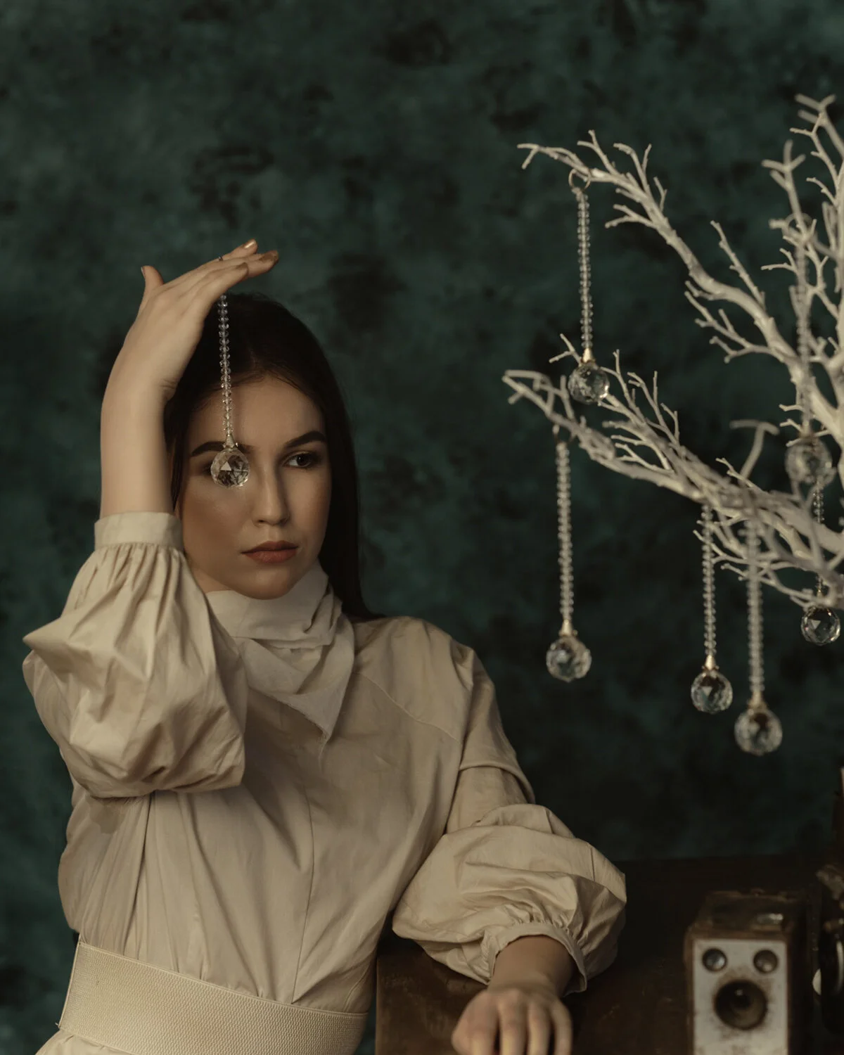 A young woman with dark hair and wearing a beige dress holds a crystal pendant hanging from a string near her forehead. She stands beside a white, leafless decorative tree decorated with similar crystal pendants and beads, against a dark green textur