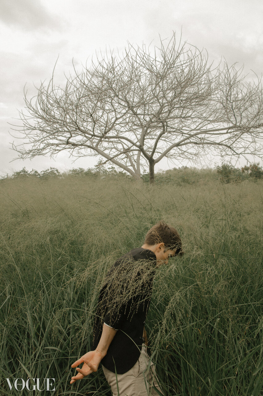 A person standing in tall grass with a leafless tree in the background, overcast sky.