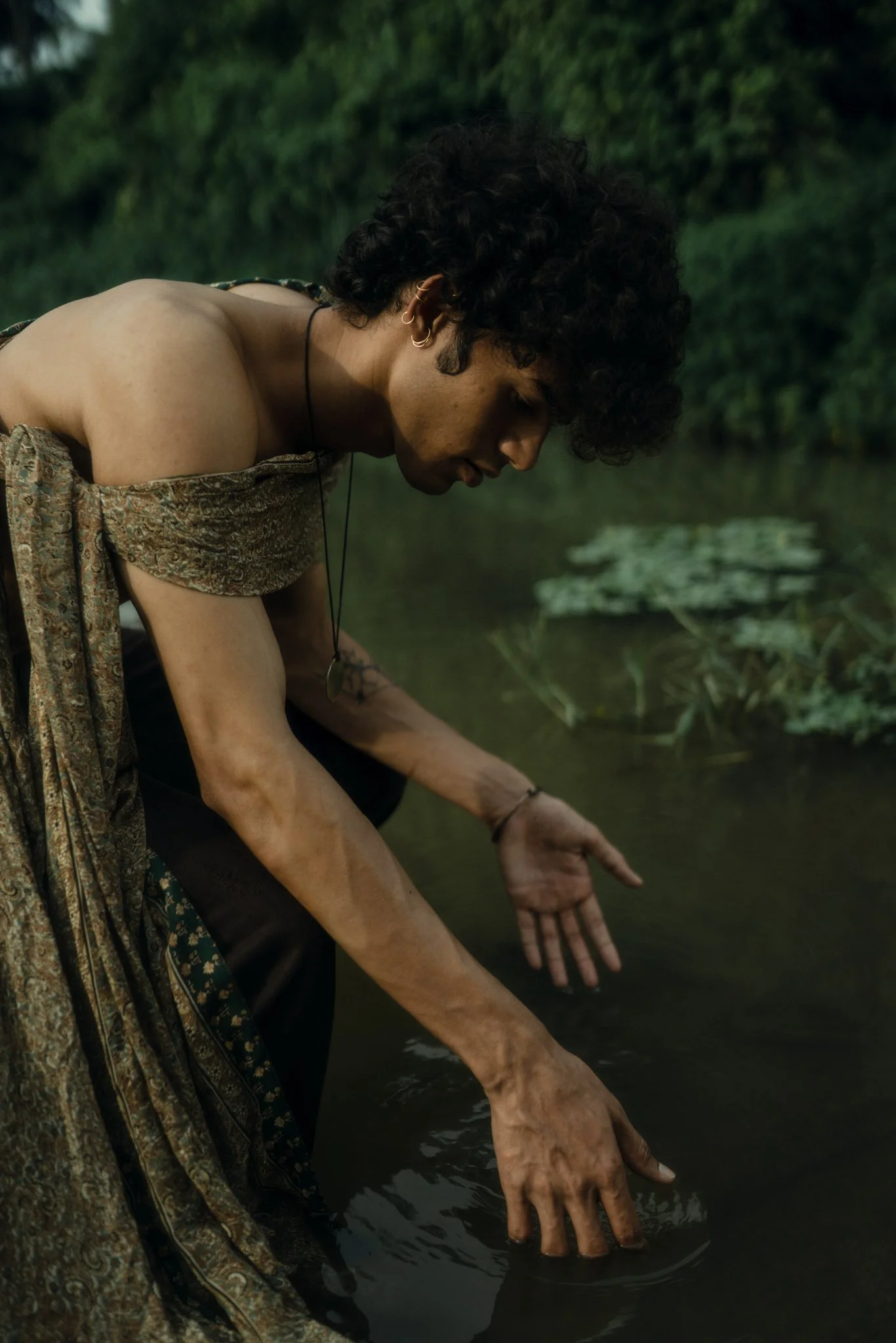 A person with curly dark hair wearing earrings and a necklace, dressed in a patterned off-the-shoulder top, leaning over a body of water with plants in the background, touching the water with their hands.