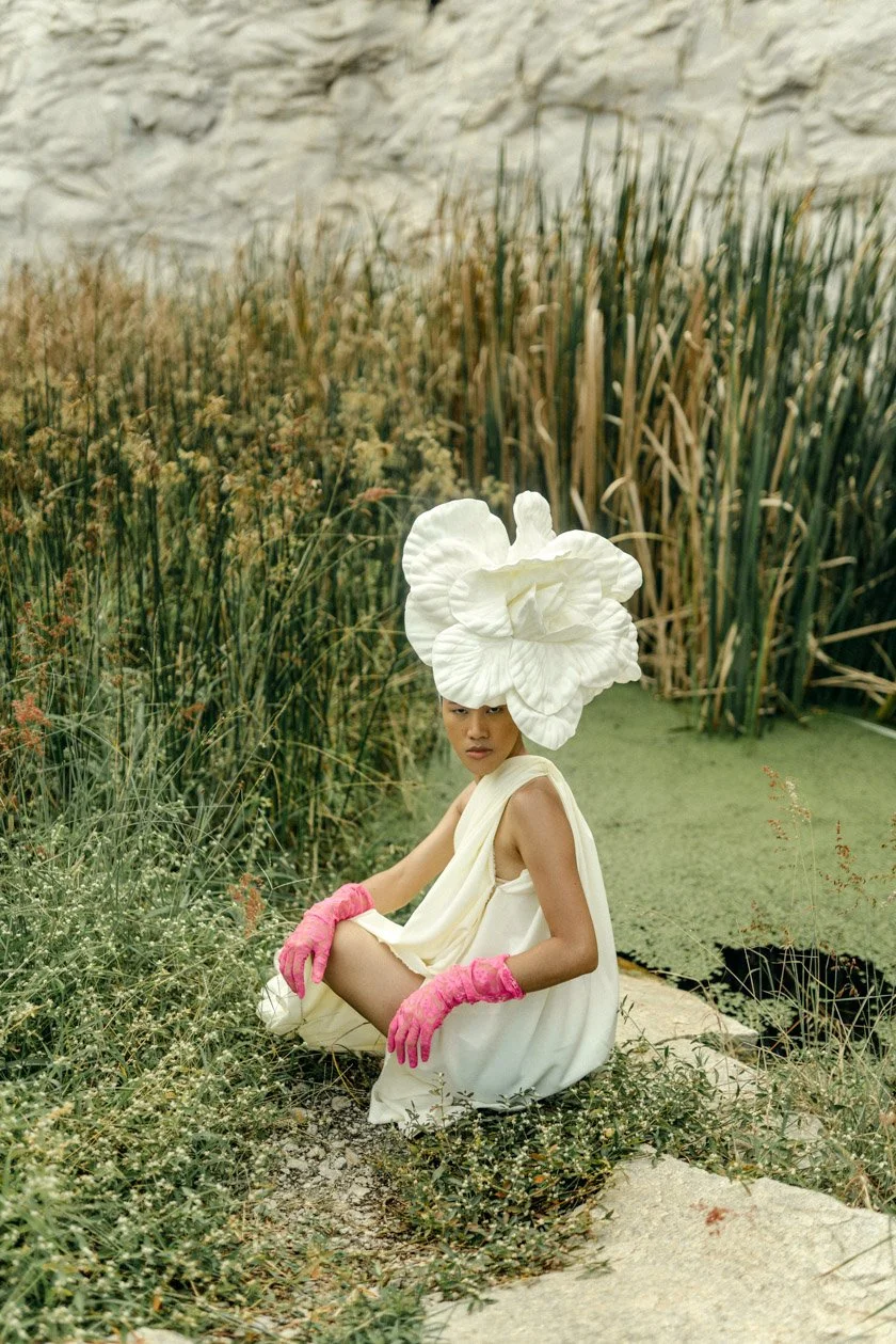 A woman wearing a large white flower hat, white dress, and pink gloves is sitting on the ground near a pond with green and brown reeds and a white rocky wall in the background.