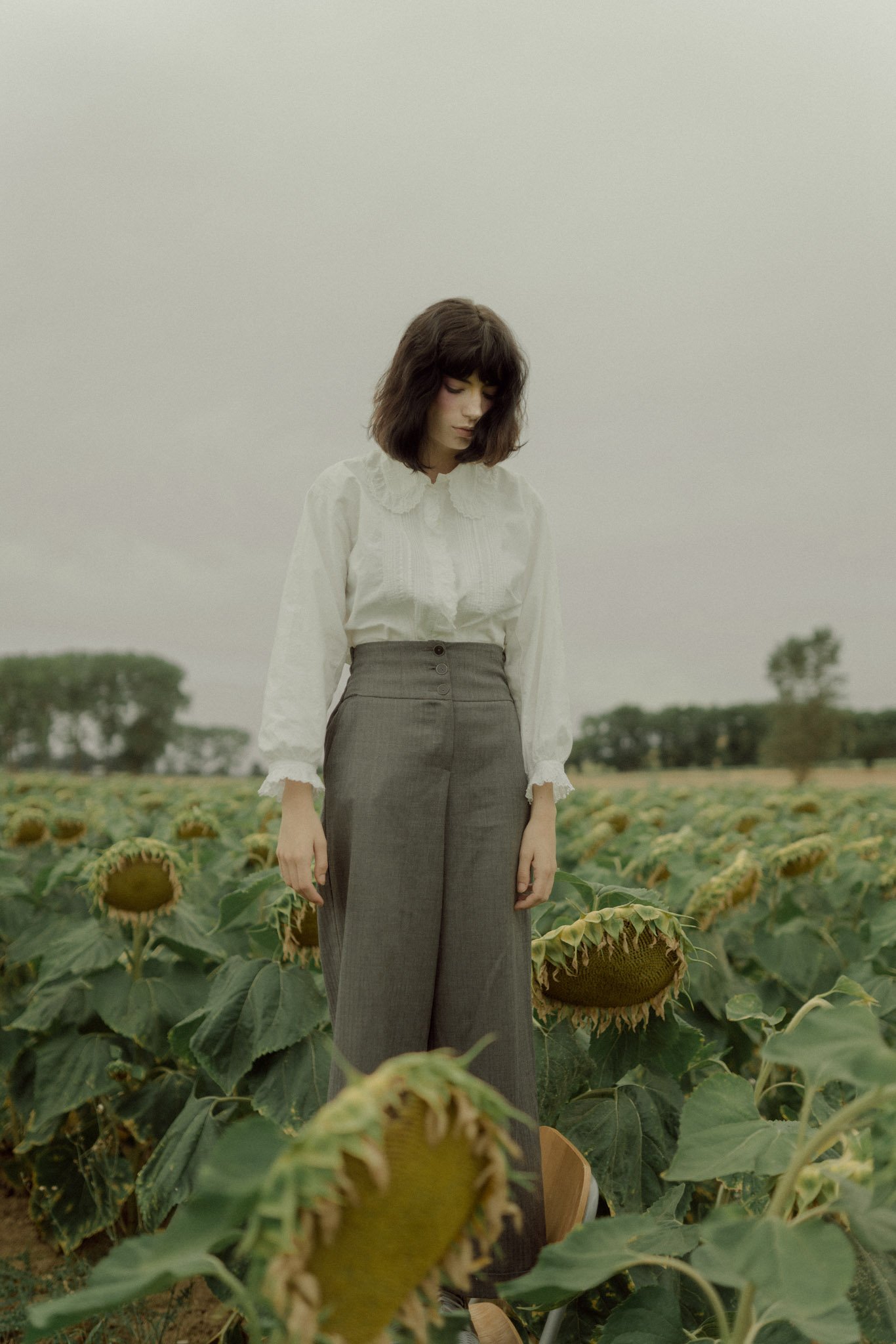 A young woman with dark hair and a white blouse standing in a sunflower field with drooping sunflowers under an overcast sky.