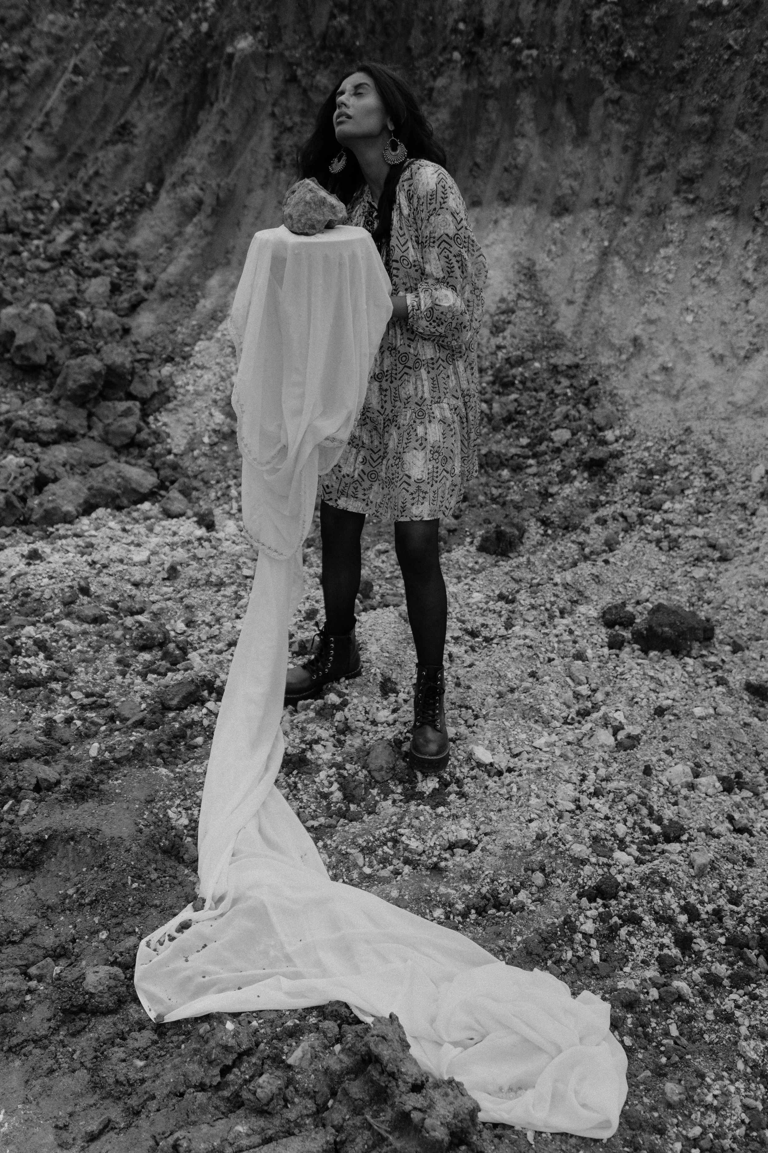 A woman stands in a rocky outdoor setting holding a white fabric draped over a pedestal with a rock on top. She wears a patterned dress, black tights, and combat boots, and has long dark hair and large earrings.