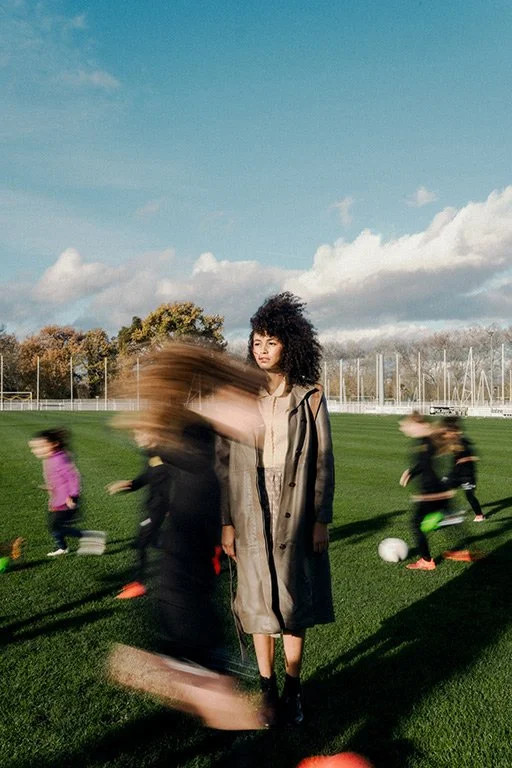Young woman standing still on a soccer field while children play soccer around her on a sunny day.