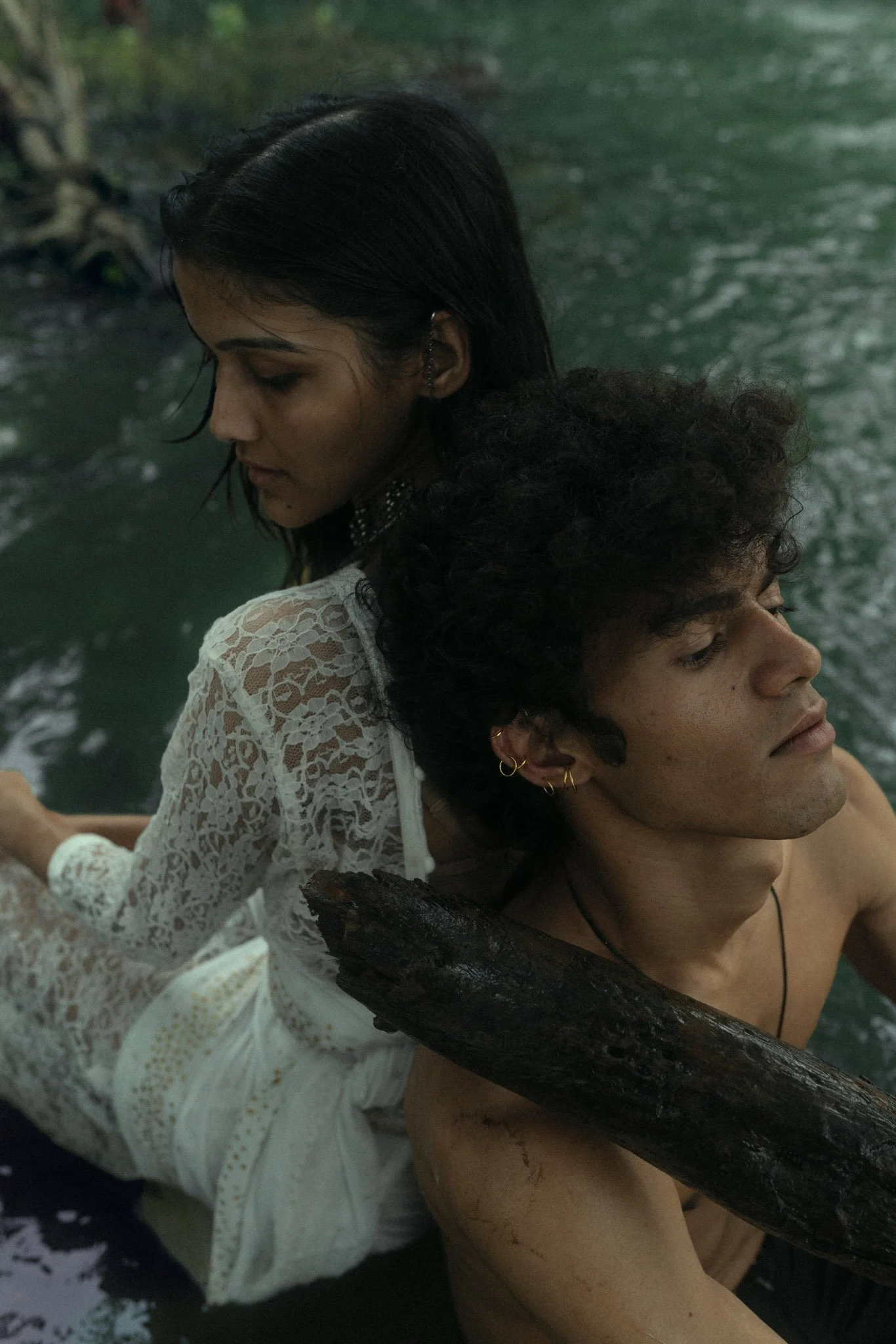 A woman in a white lace dress sitting on the back of a shirtless man in a body of water, with a river and trees in the background.