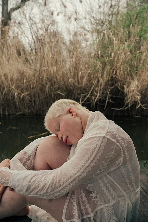 A young man with bleached blonde hair wearing a white embroidered shirt sitting outdoors by a river, resting his head on his knee with eyes closed, surrounded by tall brown reeds and trees.