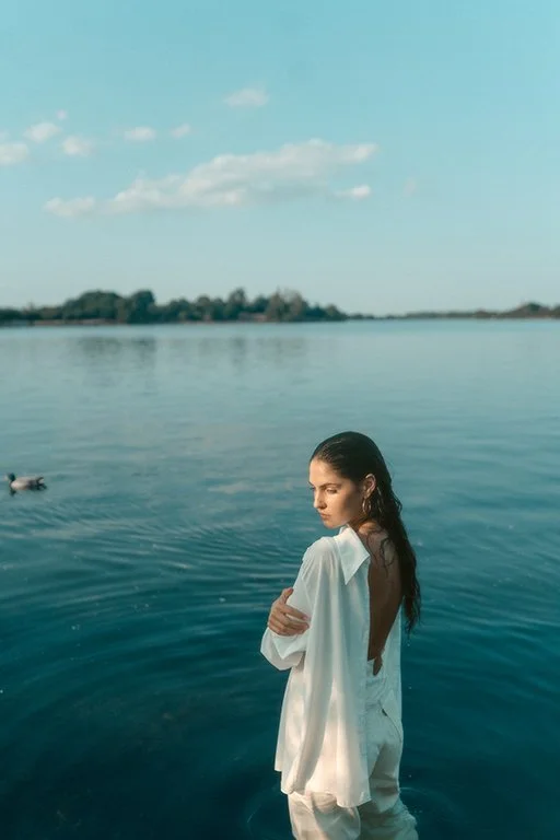 A young woman stands in a large body of water, wearing a white dress with an open back, looking to the side with a serene expression. Ducks are swimming nearby, and the sky is partly cloudy with a distant shoreline visible.