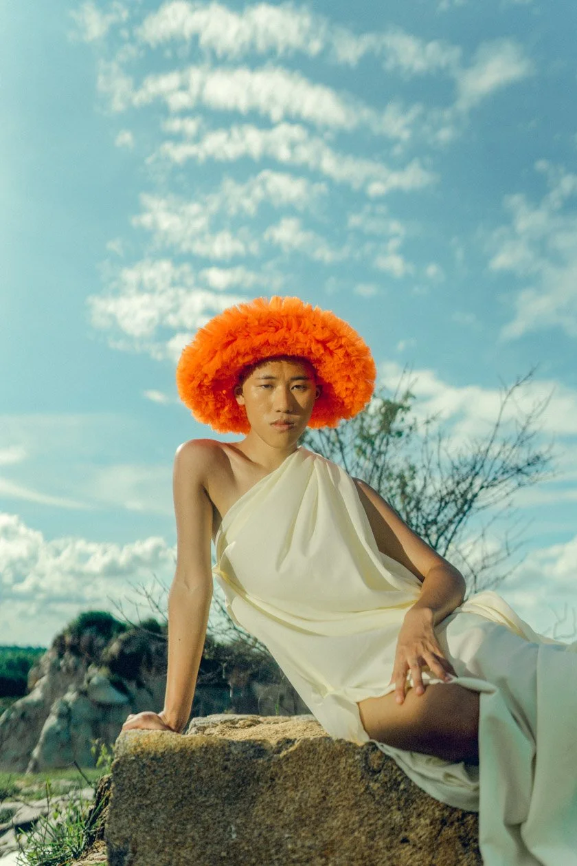Person wearing an orange fuzzy hat and a white draped dress, sitting outdoors on a rock with a background of sky and clouds.