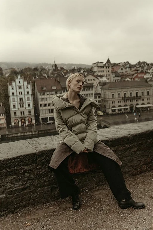 A woman sitting on a stone ledge overlooking a cityscape with old buildings on a cloudy day.