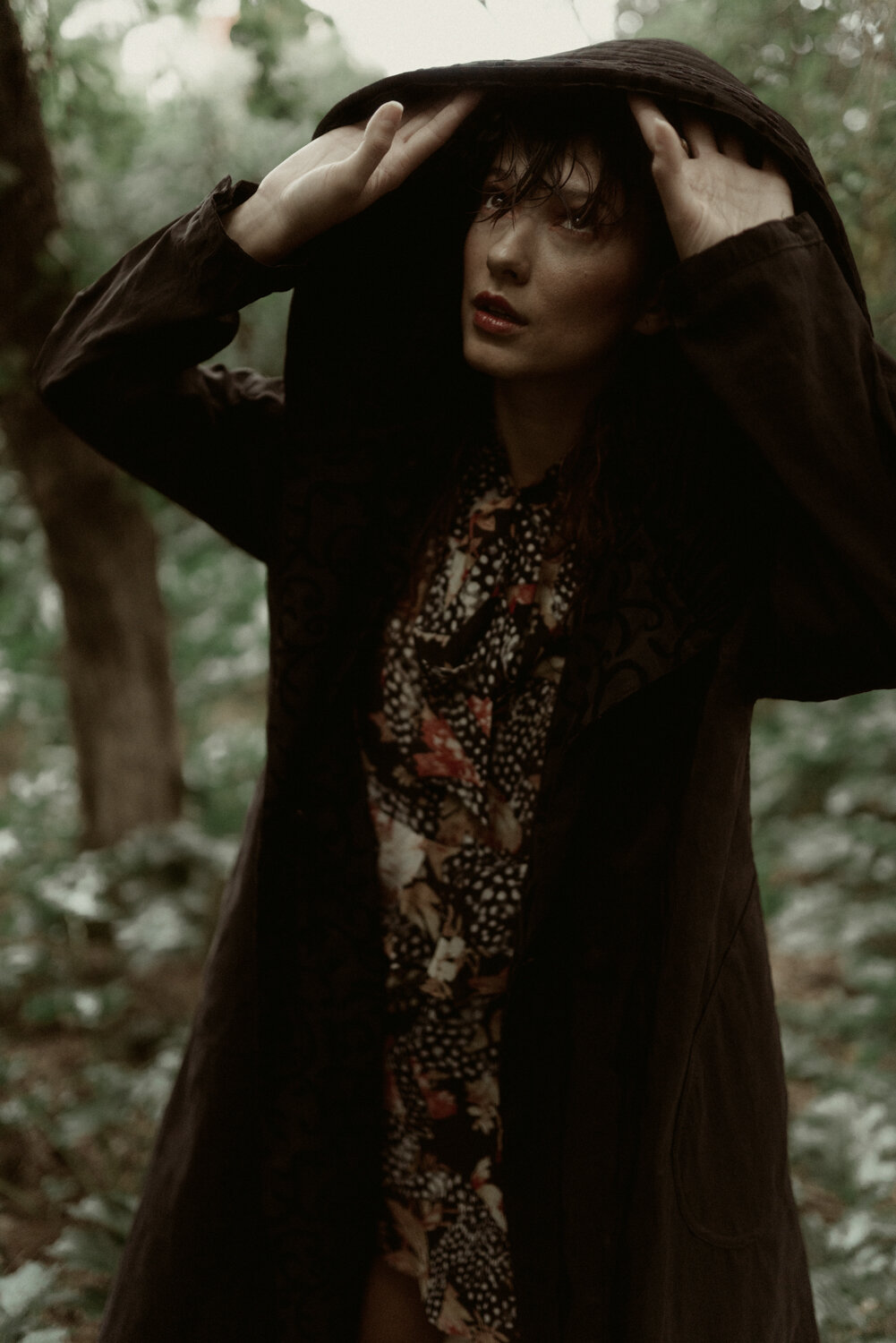 A woman in a dark coat and patterned dress standing outdoors in a wooded area, holding a hood over her head and gazing into the distance.