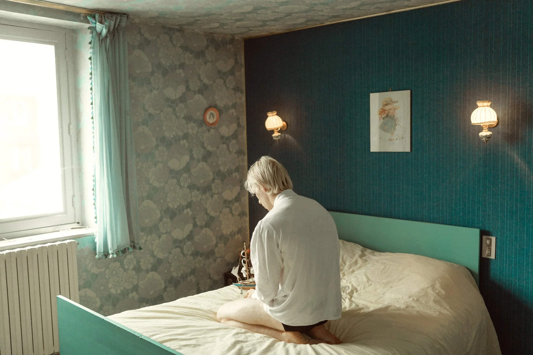 A person with light hair kneeling on a bed in a bedroom, looking at a ship model. The room has blue and floral wallpaper, a large window with curtains, and two wall-mounted lamps.