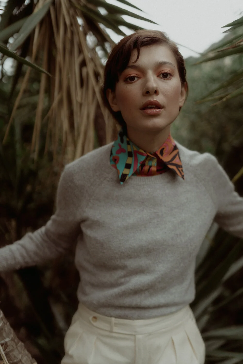 A woman with short brown hair and light skin standing outdoors among tall, dry plants, wearing a light gray sweater with a colorful patterned scarf around her neck.