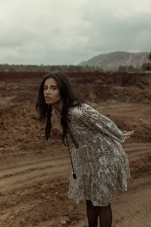 A woman with dark, wavy hair wearing a patterned dress and earrings is outdoors in a barren, muddy landscape with a distant mountain in the background.