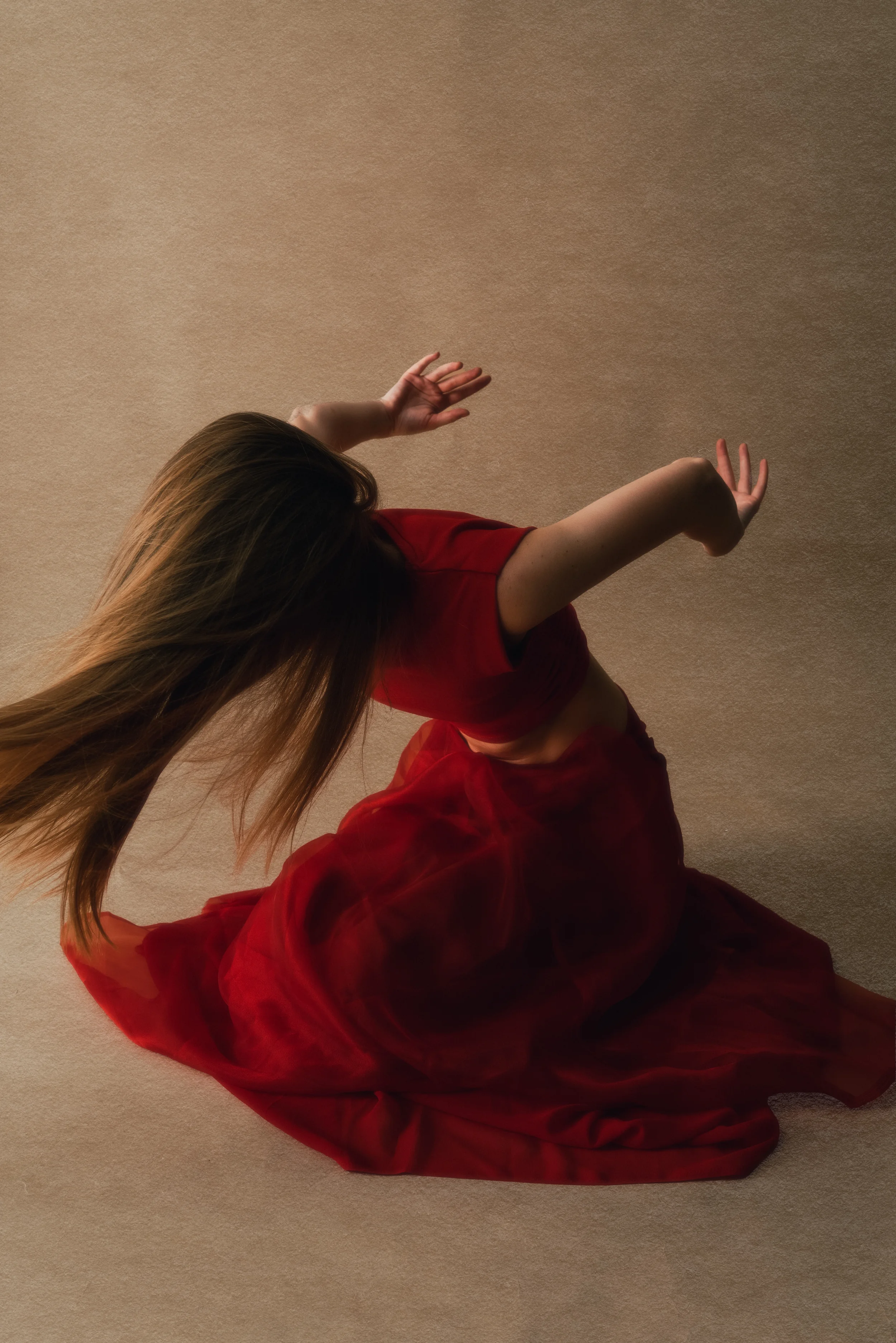 A woman with long brown hair wearing a red dress sitting on the floor with a beige carpet and a beige wall in the background.