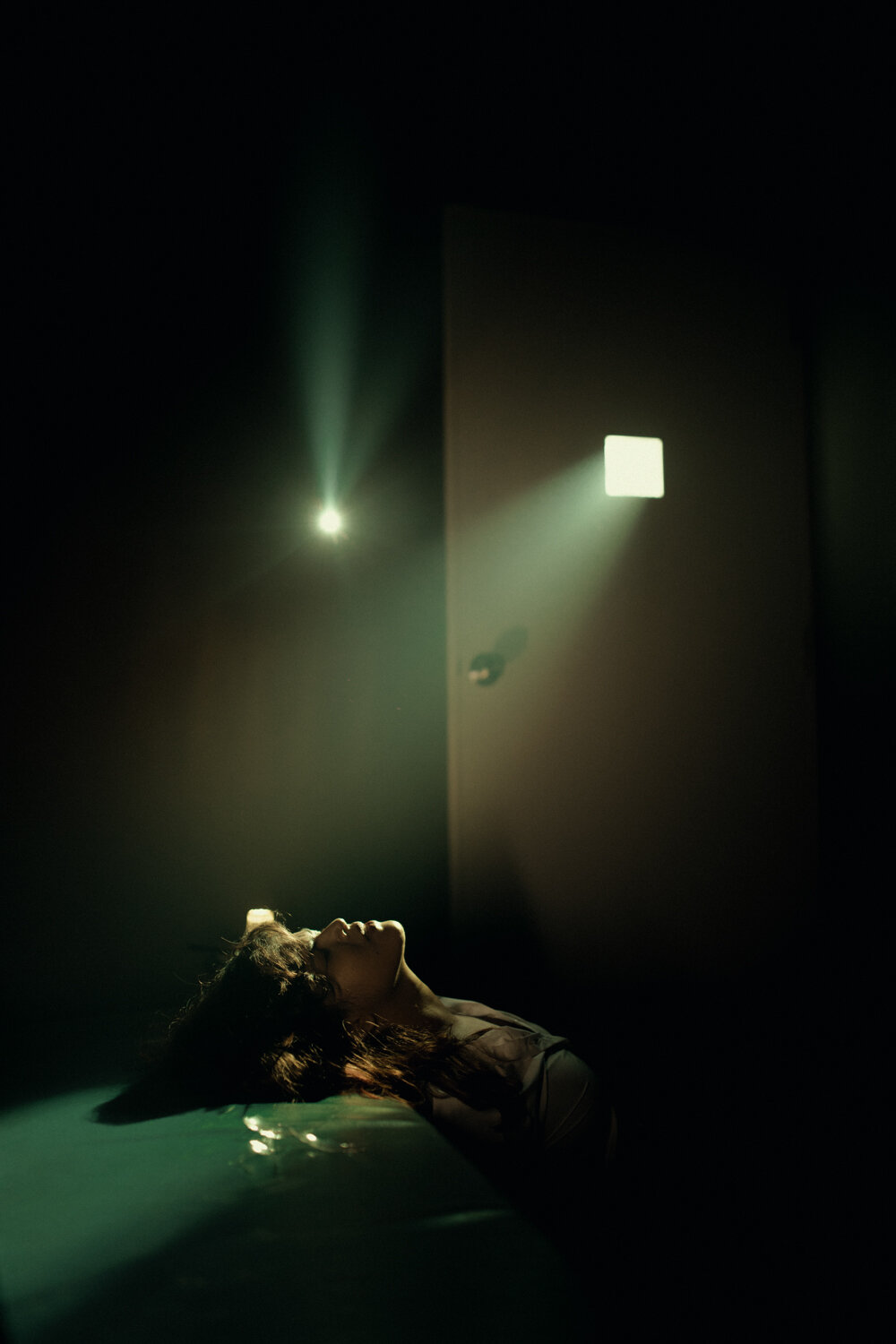A woman lying on the floor in a dark room, illuminated by a small light coming through an open door, with light rays creating a dramatic atmosphere.