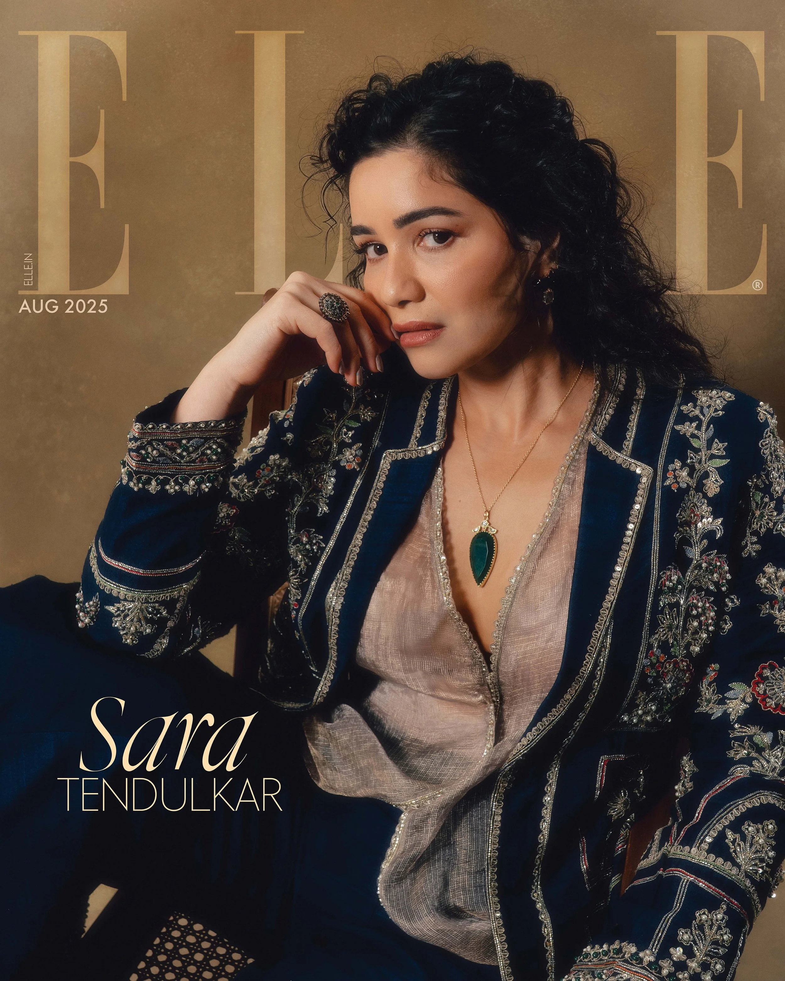 A woman with dark curly hair wearing a navy blue embroidered jacket, a beige blouse, and jewelry including a large ring, earrings, and a gold necklace with a green pendant, sitting against a beige background on the cover of Elle magazine, August 2025