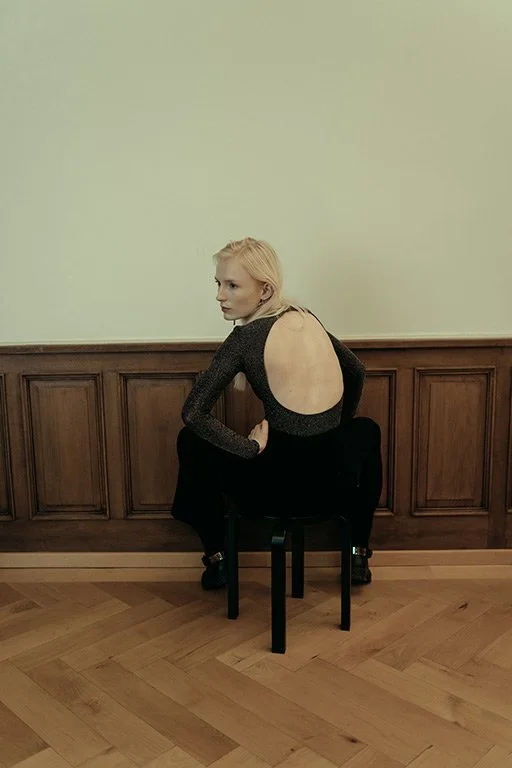A woman with blonde hair and light skin sitting on a black chair against a wooden-paneled wall, wearing a black dress with a large circular cutout on the back.