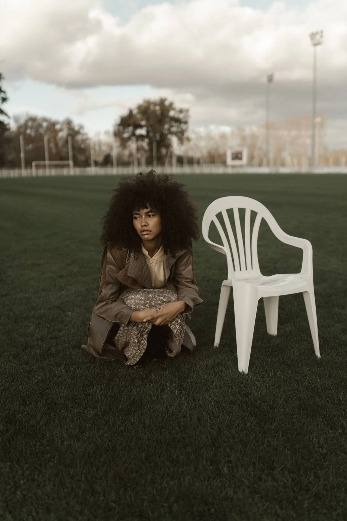 A woman with curly hair squatting on grass next to a white plastic chair, with a cloudy sky and a football field in the background.
