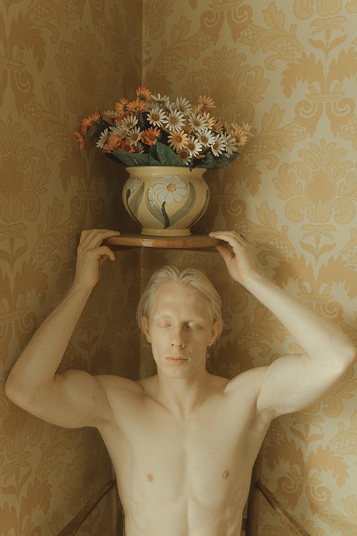 A shirtless person with short blond hair balancing a large vase of flowers on a wooden plank above their head inside a room with patterned wallpaper.