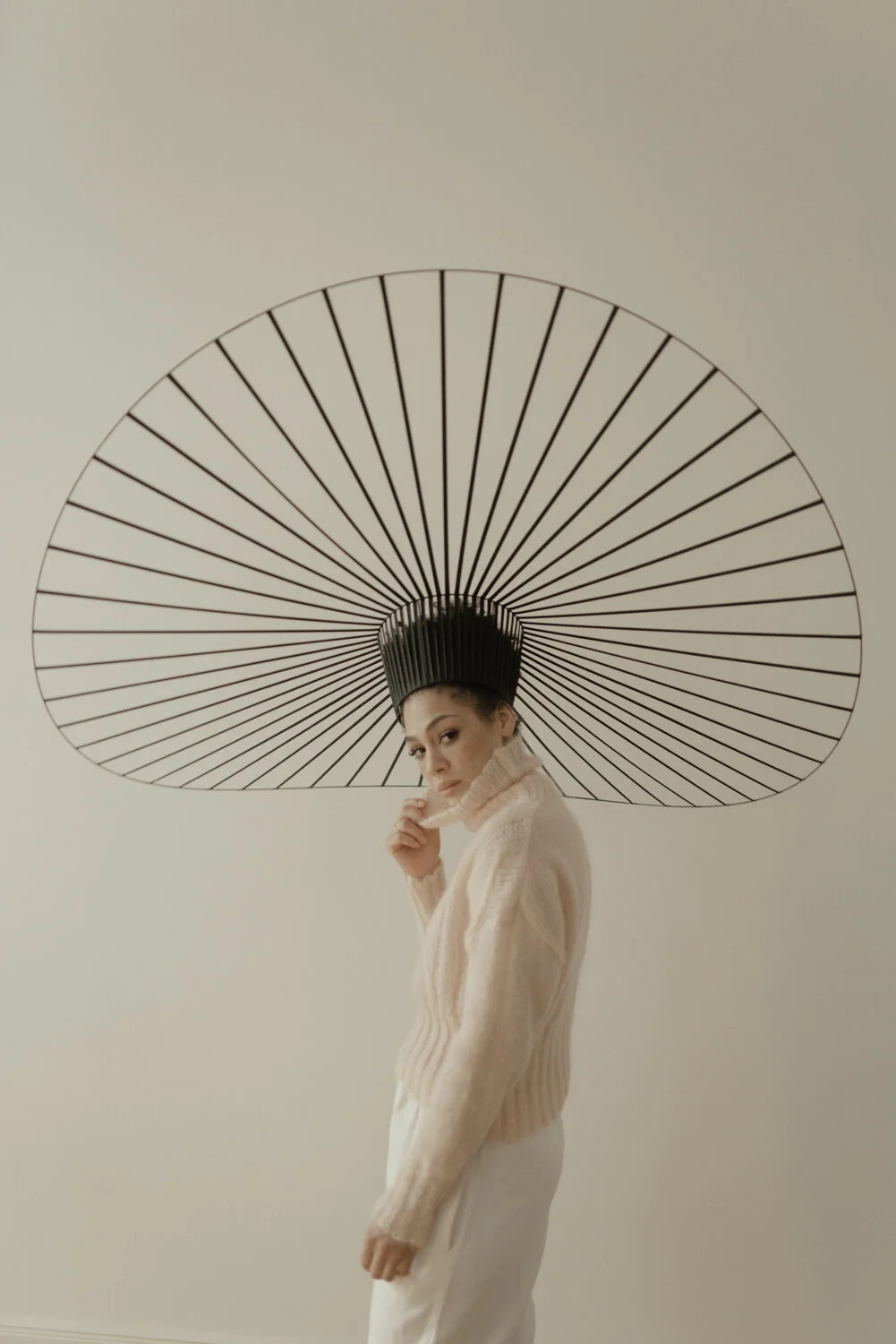 A woman wearing a cream turtleneck sweater and white pants posing in front of a minimalist art piece resembling an umbrella with black spokes on a beige wall.