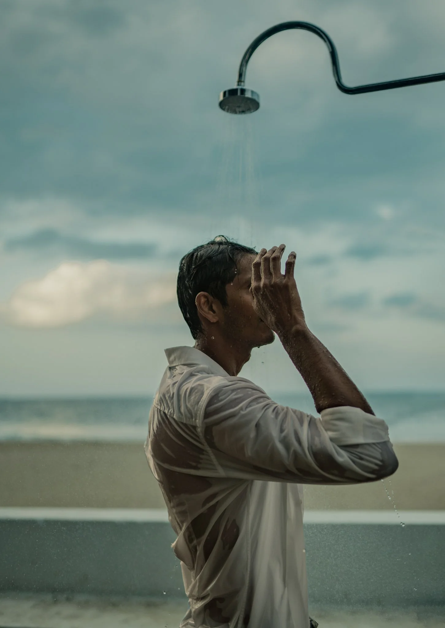 A man under an outdoor shower on a beach, he is rinsing off with water, wearing a wet shirt, with a cloudy sky in the background.