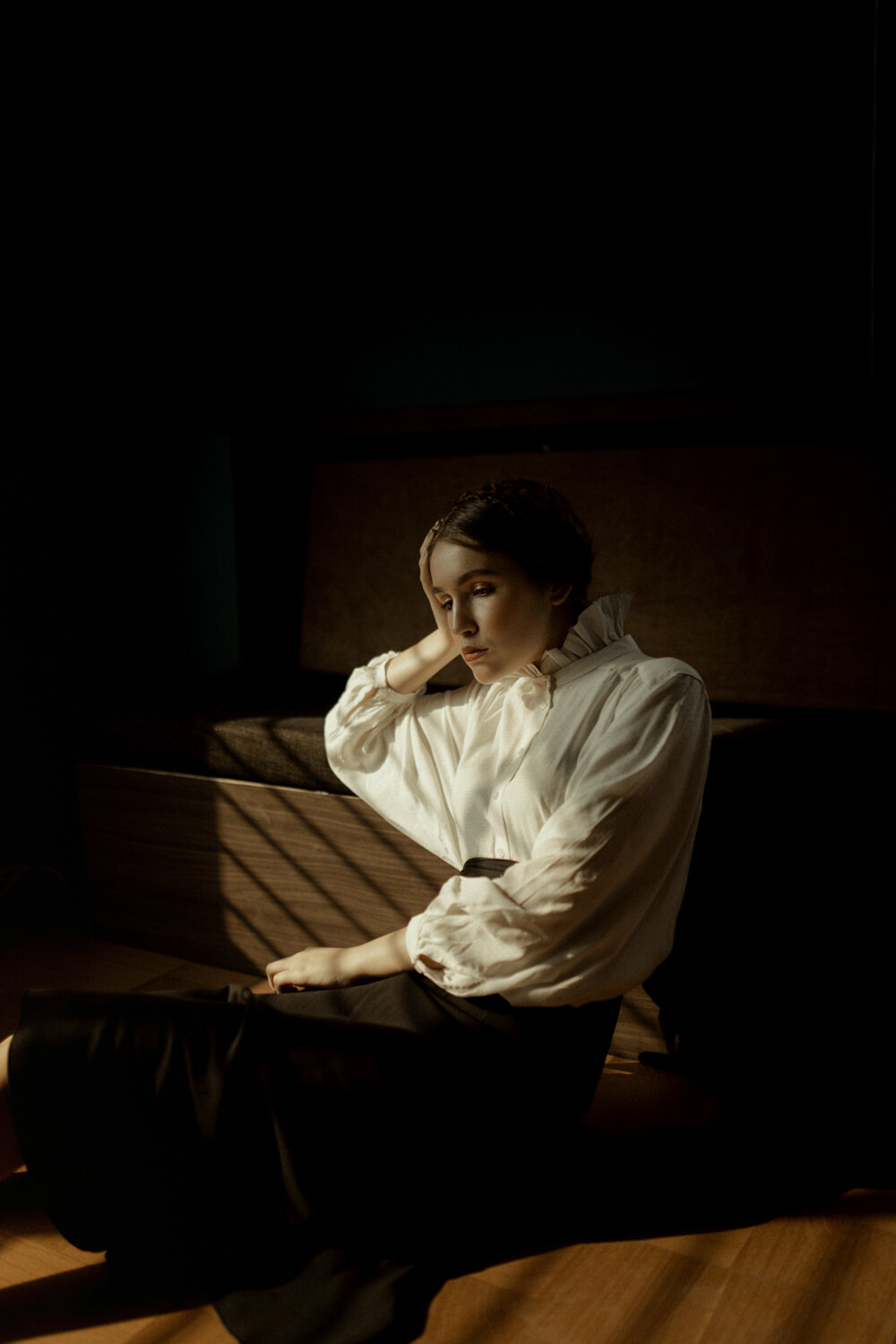 A woman with short hair and wearing a white blouse, sitting on the floor with one hand resting on her head, in a dimly lit room with soft light creating shadows.