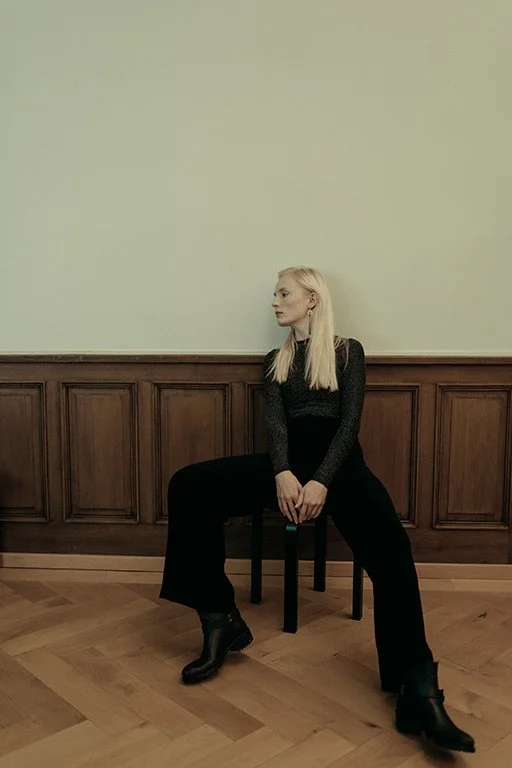 A blonde woman in black clothing and boots sits on a chair with her legs apart, against a wall with wooden paneling in a room with wooden flooring.