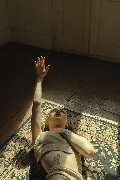 Child lying on a patterned rug on the floor, reaching upwards with one arm, with sunlight illuminating part of her face and body.