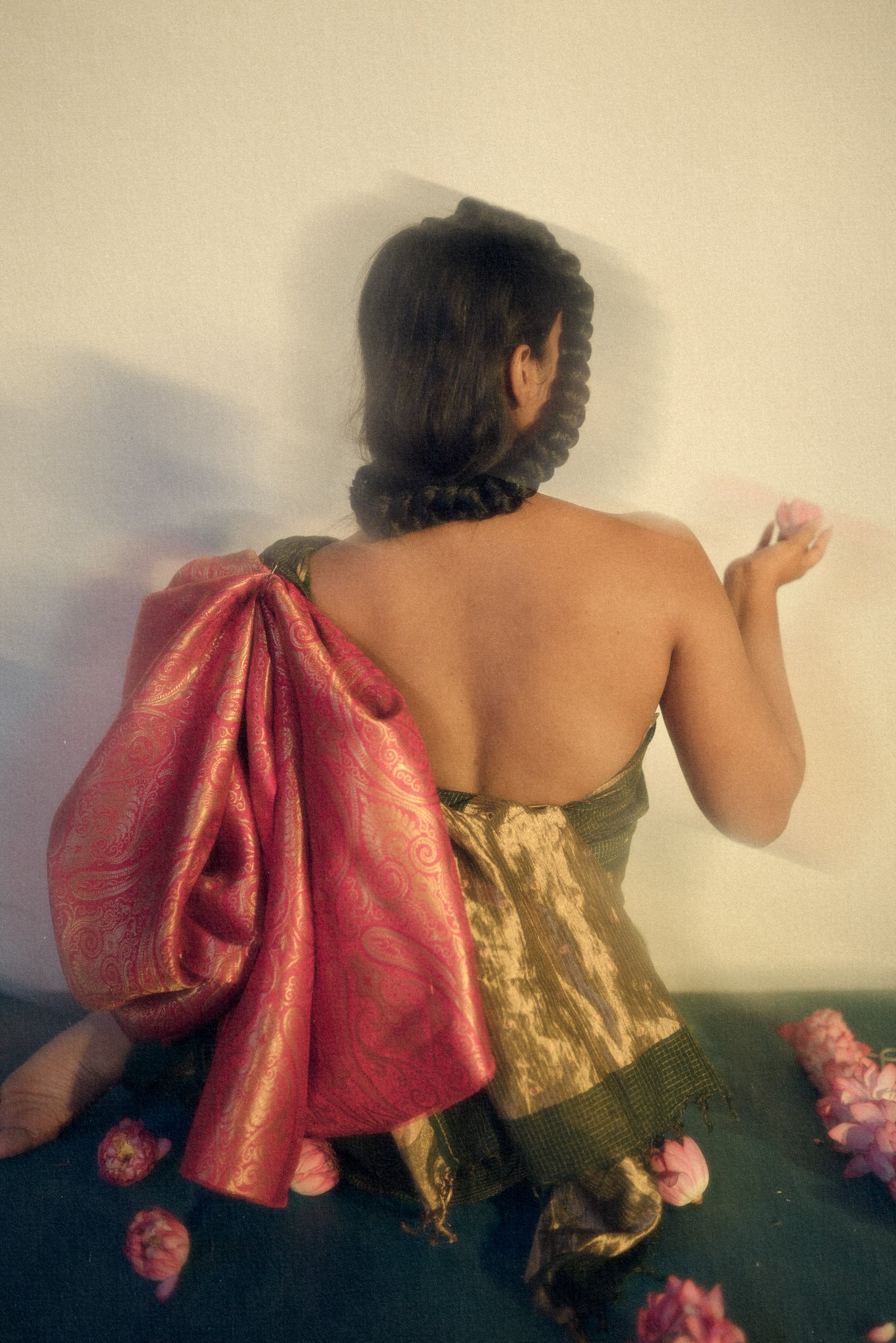 A woman with an elaborate braided hairstyle, wearing traditional clothing with a large red and gold sash, sitting on a dark surface with pink flower petals, facing away from the camera.
