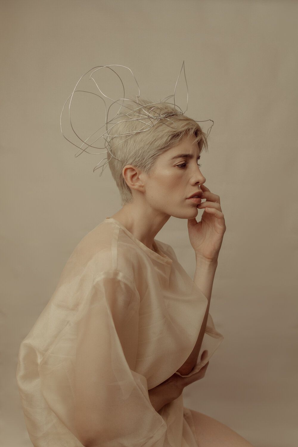 A woman with platinum blonde hair styled short, wearing a sheer, beige top, and holding her chin thoughtfully. She has a wire sculpture on her head resembling abstract ears or a headpiece.