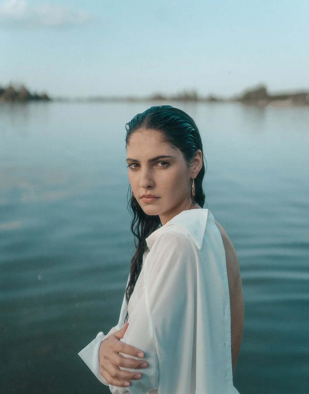 A woman with dark, wet hair and hoop earrings standing near a calm body of water, wearing a white shirt with the back open.