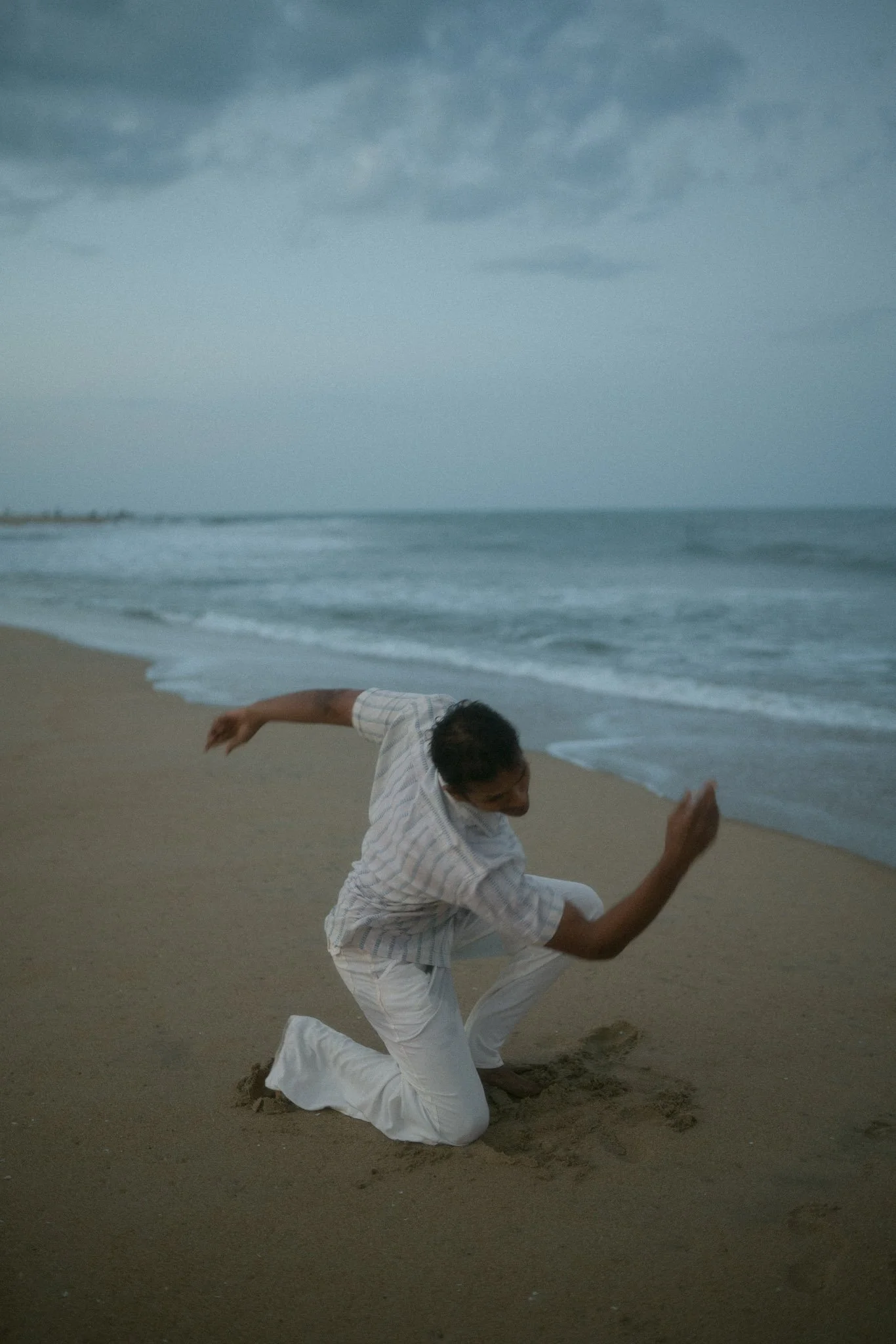 A man kneeling on a sandy beach near the ocean, with dark clouds in the sky, during early evening or dusk.