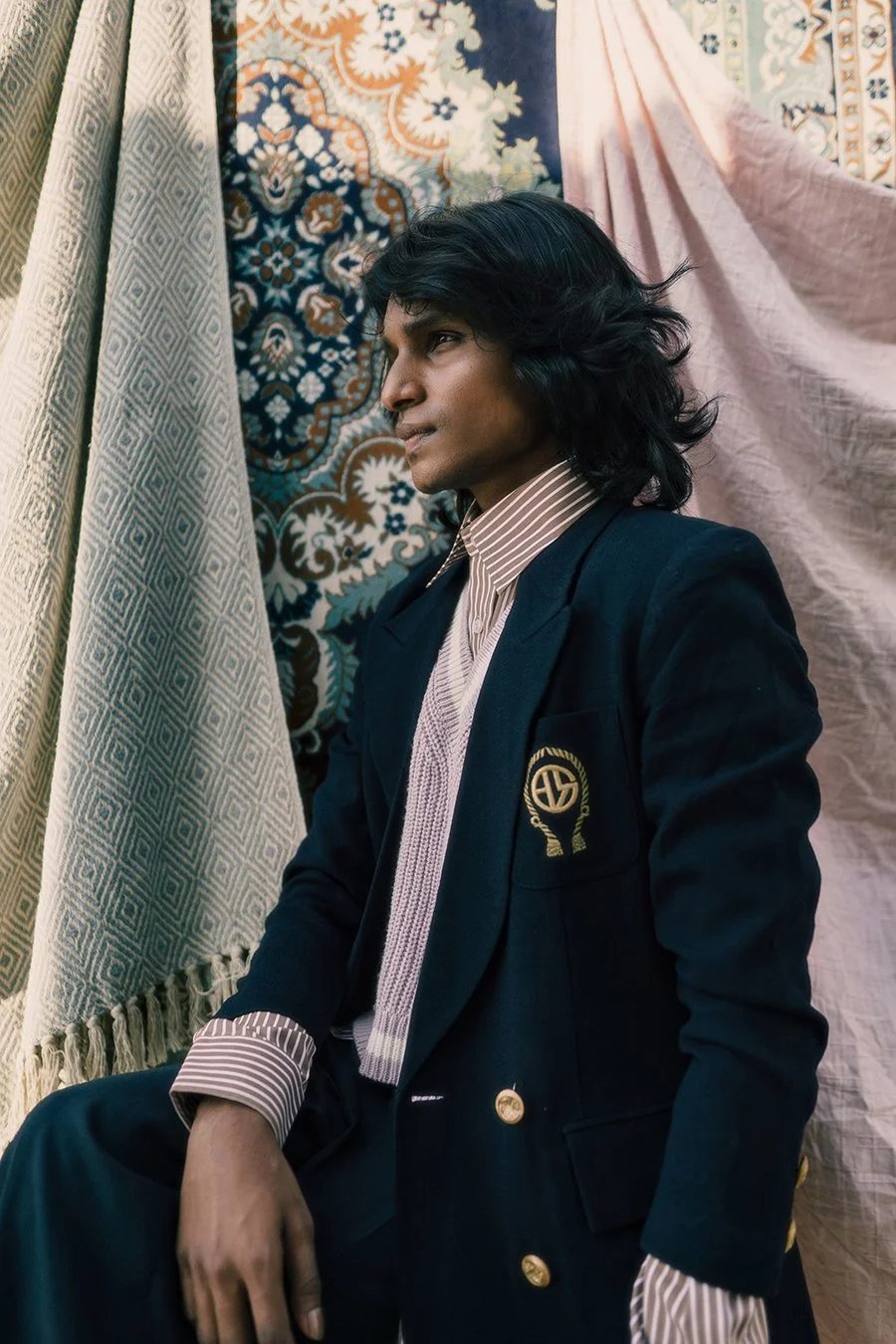 A person with dark, wavy hair wearing a black blazer with a school emblem on the pocket, a striped shirt, and a light-colored sweater, sitting in front of patterned curtains and textiles.