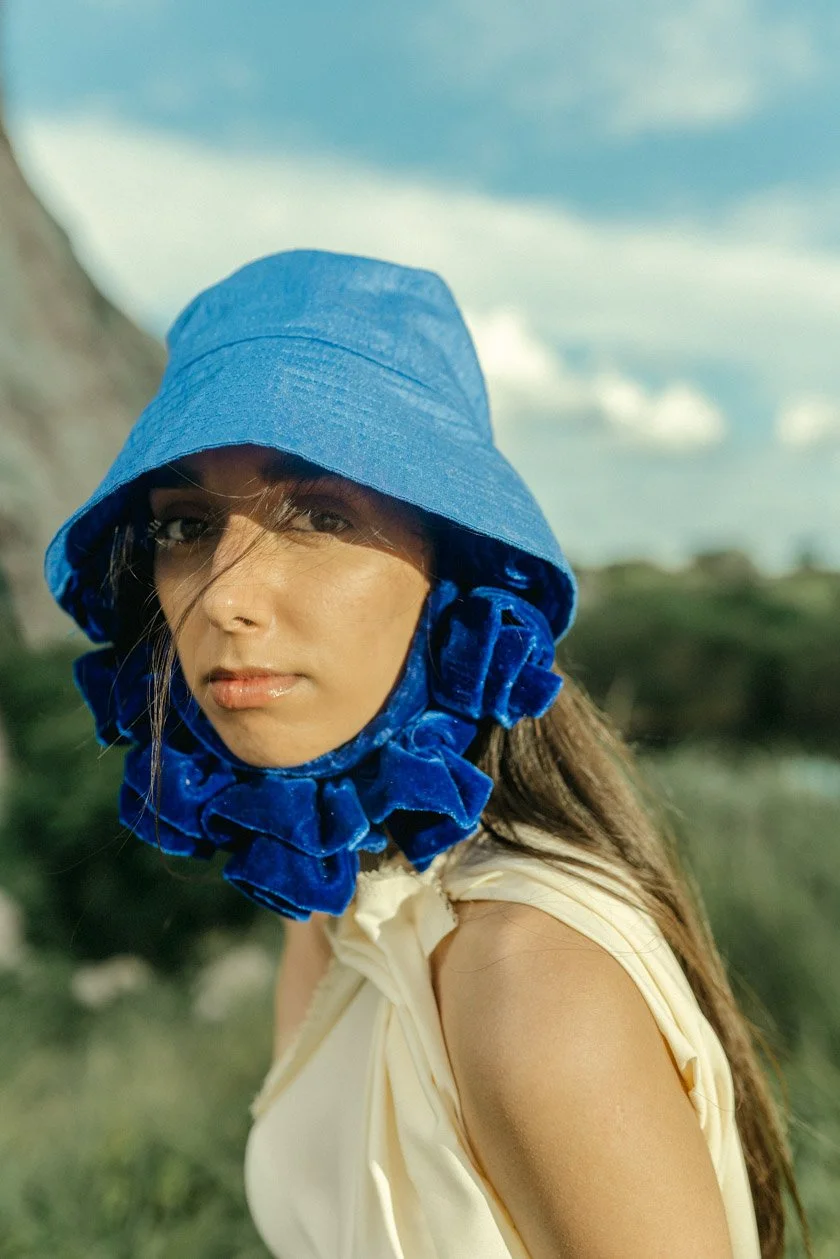 A woman outdoors wearing a blue wide-brimmed hat with a ruffled fabric around her neck, and a cream-colored sleeveless top, with a blurred natural landscape and cloudy sky in the background.