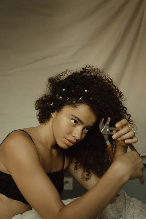 A woman with curly hair with pearl accents, wearing a black strap top, looks focused while using pliers on her hair.