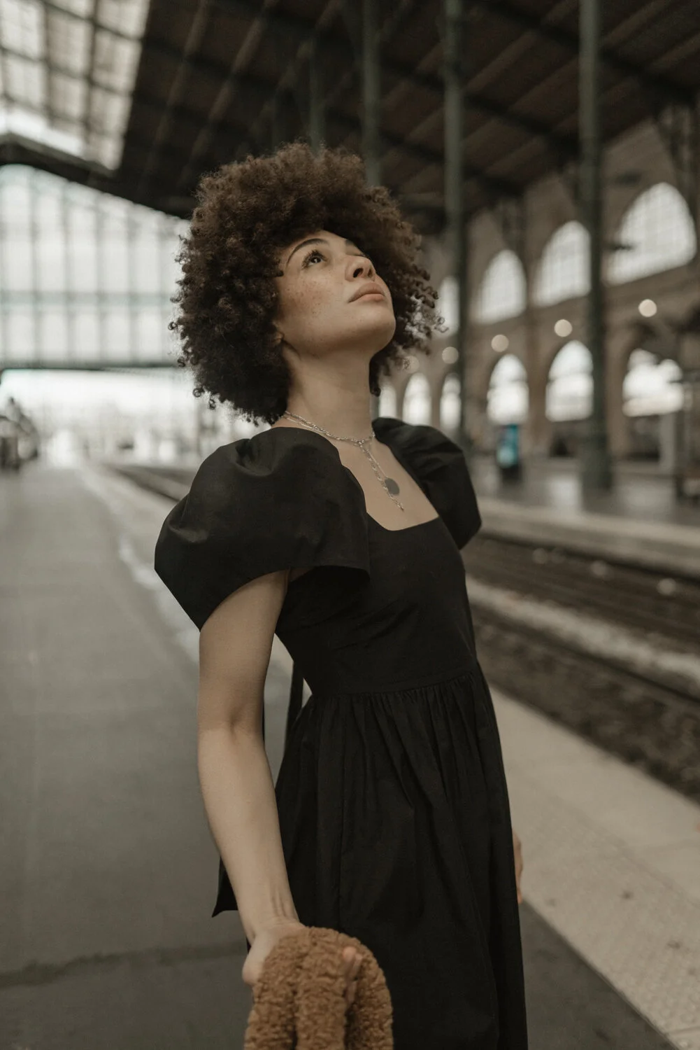 A woman with curly hair wearing a black dress and necklace standing on a train platform, looking upward.