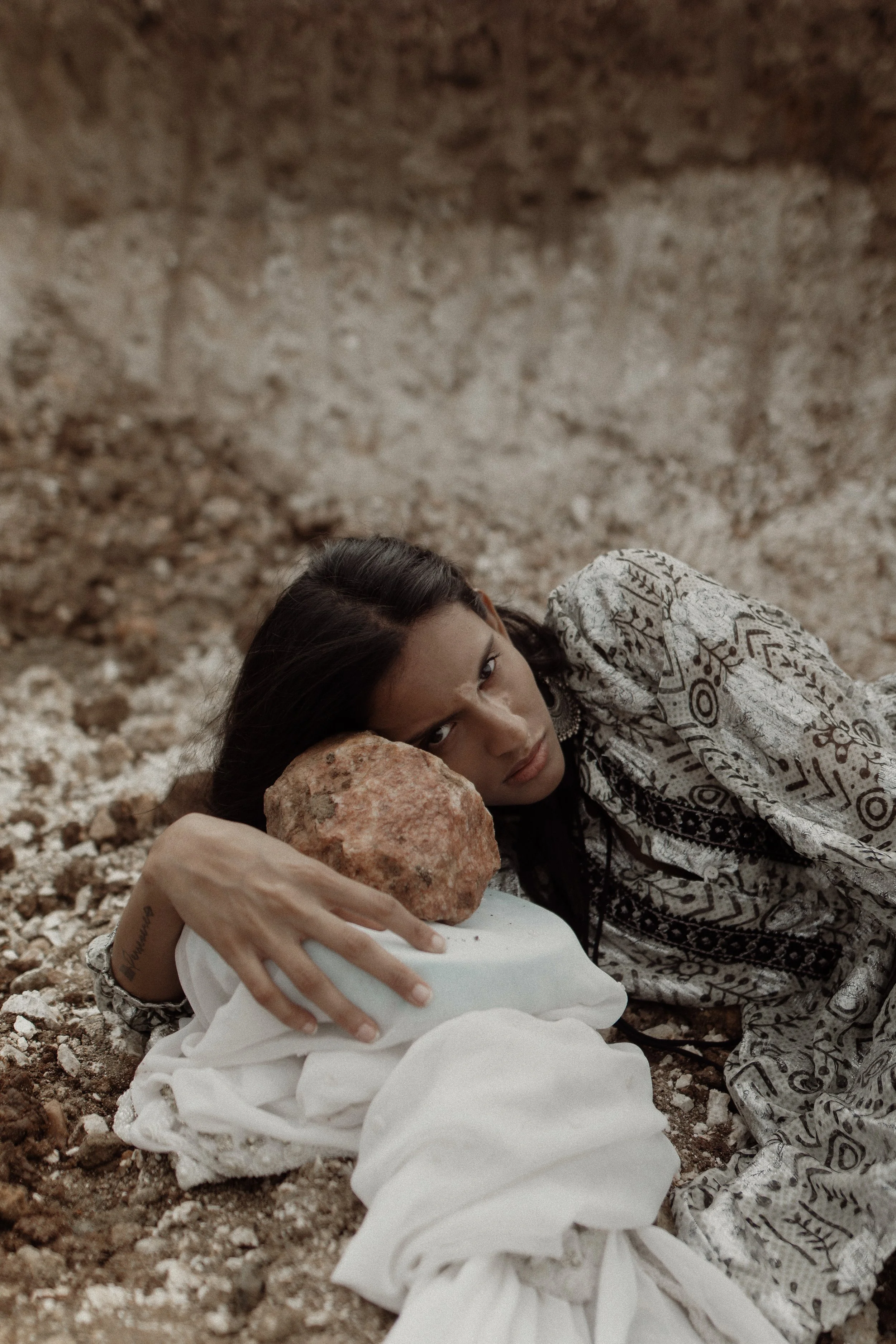 A woman lying on rocky ground holding a large rock against her head, wearing a patterned shirt with a serious expression, with a blank background.