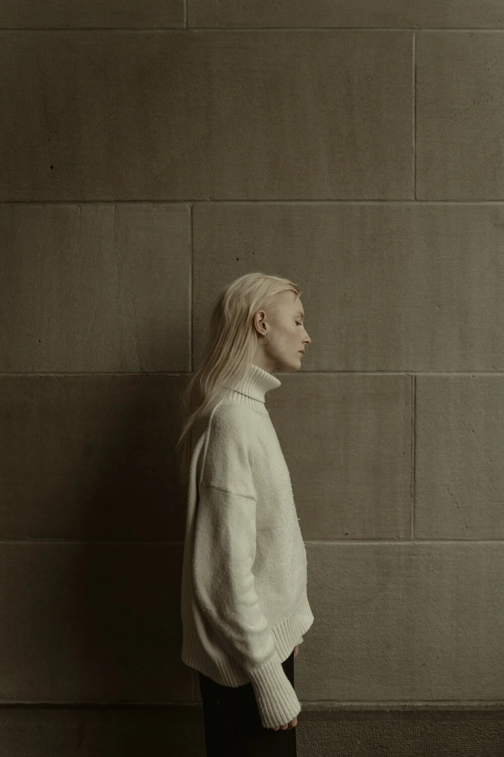 Side profile of a woman with platinum blonde hair standing against a plain, textured wall, wearing a cream-colored turtleneck sweater.