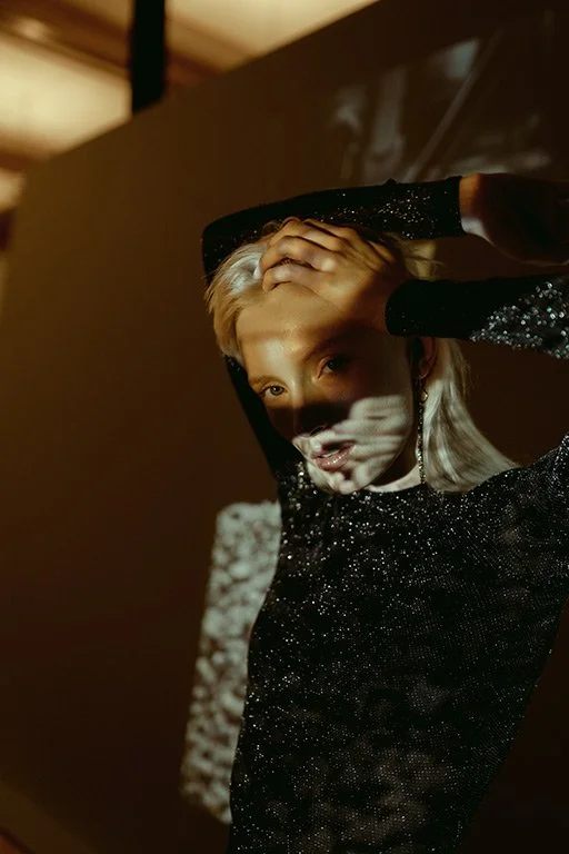 A woman with platinum blonde hair wears a black, sparkly top and holds her head with one hand, looking at the camera with a soft expression. A shadow cast on her face creates a patterned light effect.