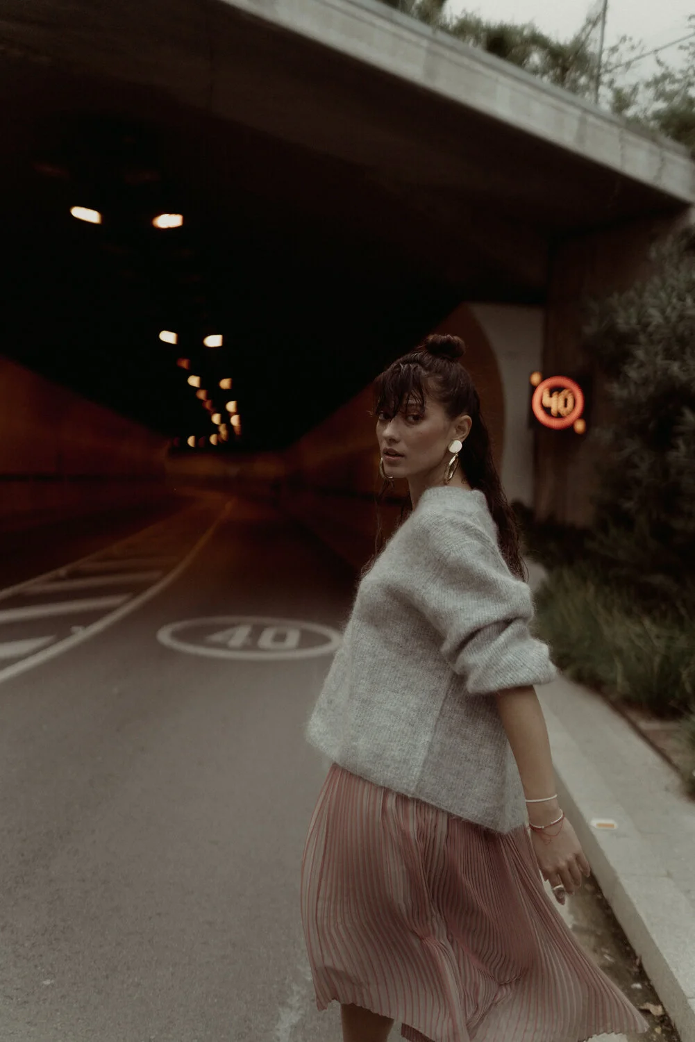 A young woman with dark hair styled in a bun, wearing a gray sweater and a pleated pink skirt, stands on a city street near a tunnel with a speed limit sign of 40 mph. She is looking over her shoulder, accessorized with earrings and bracelets, with a