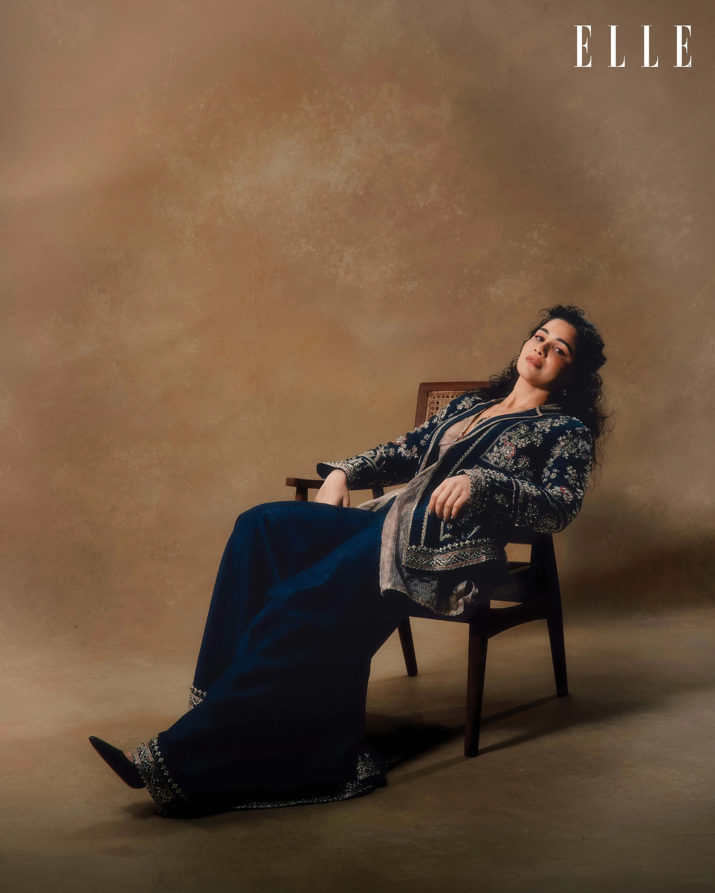A woman with curly dark hair sitting on a wooden chair against a beige background, wearing a dark embroidered jacket and matching wide-leg pants, posing confidently for a fashion photoshoot for Elle magazine.