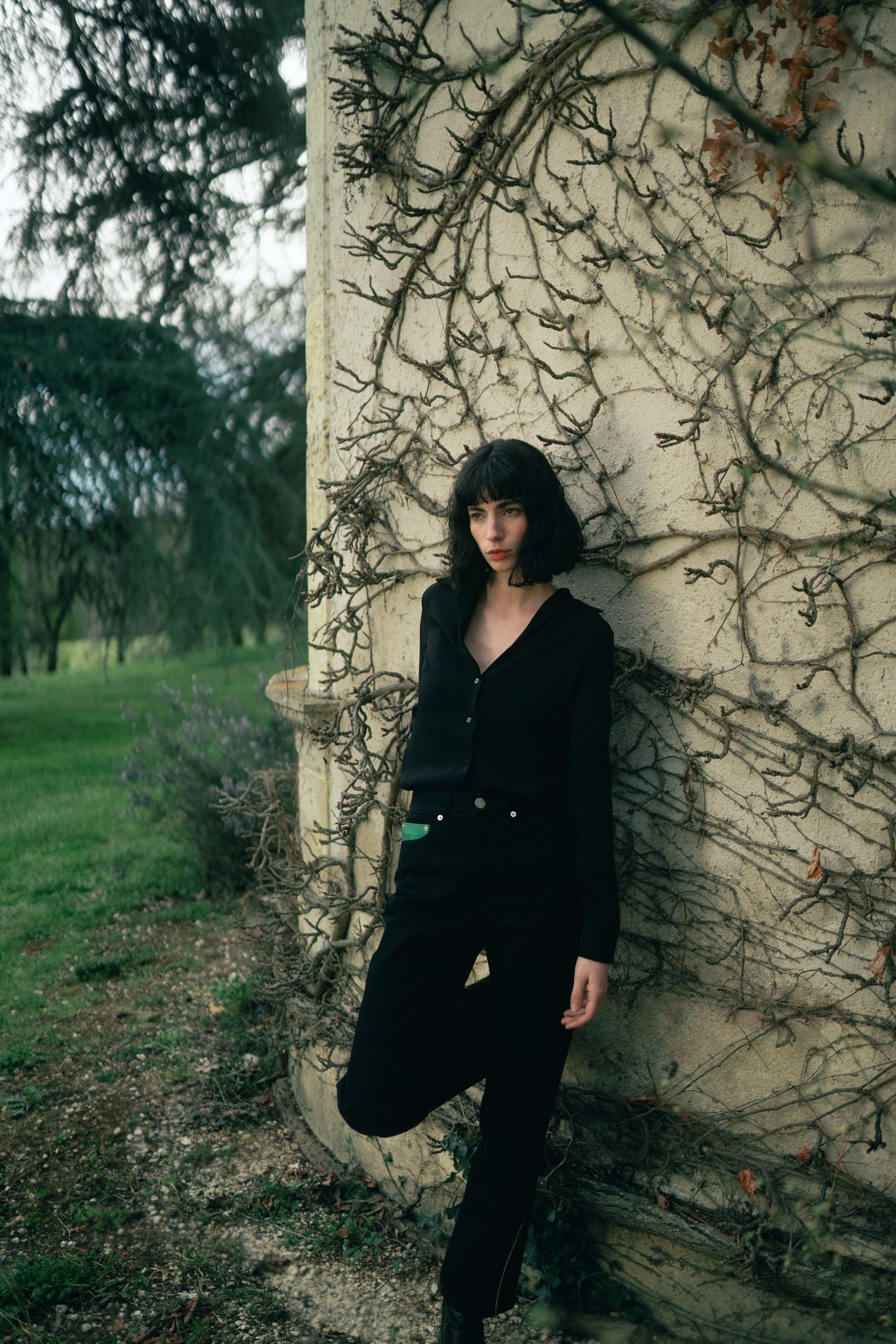 A woman with black hair and bangs standing outdoors against an ivy-covered wall, wearing a black shirt and pants, with trees and greenery in the background.