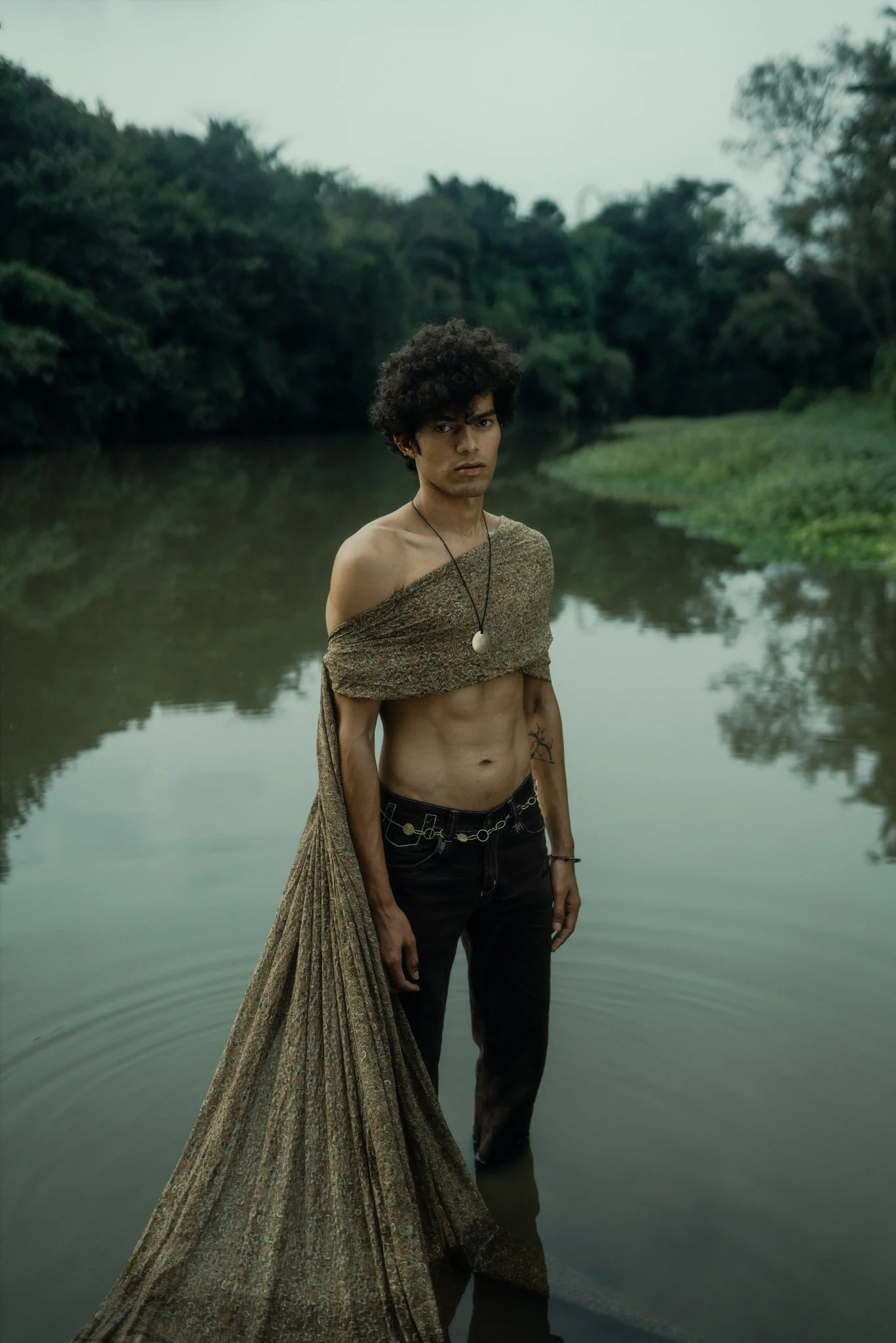 A young man with short curly hair, wearing dark pants and a chain belt, stands in a body of water, partially draped in a patterned fabric around his shoulders, with a forested background and overcast sky.