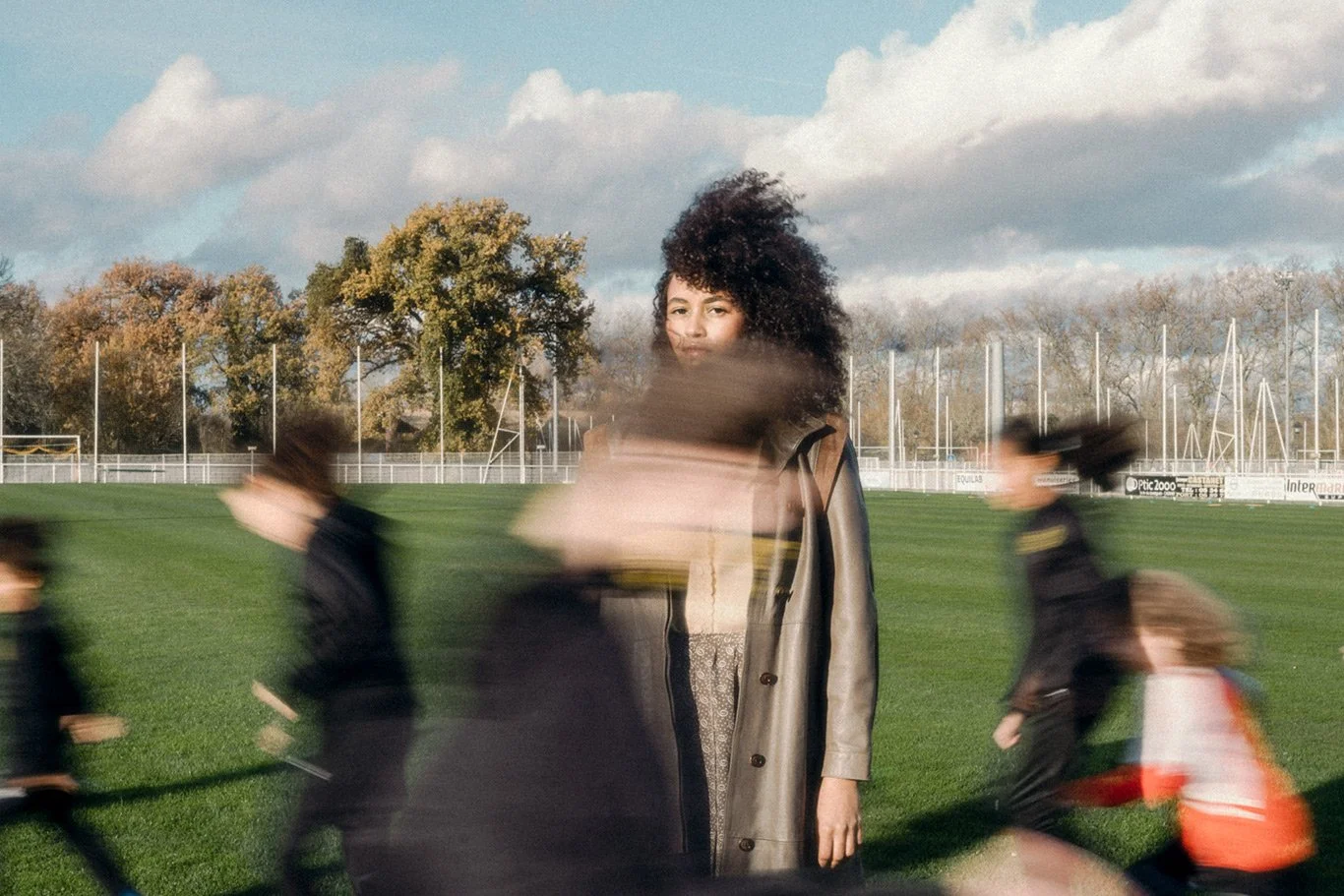 A woman with curly hair standing on a grassy field with blurred children running past her.